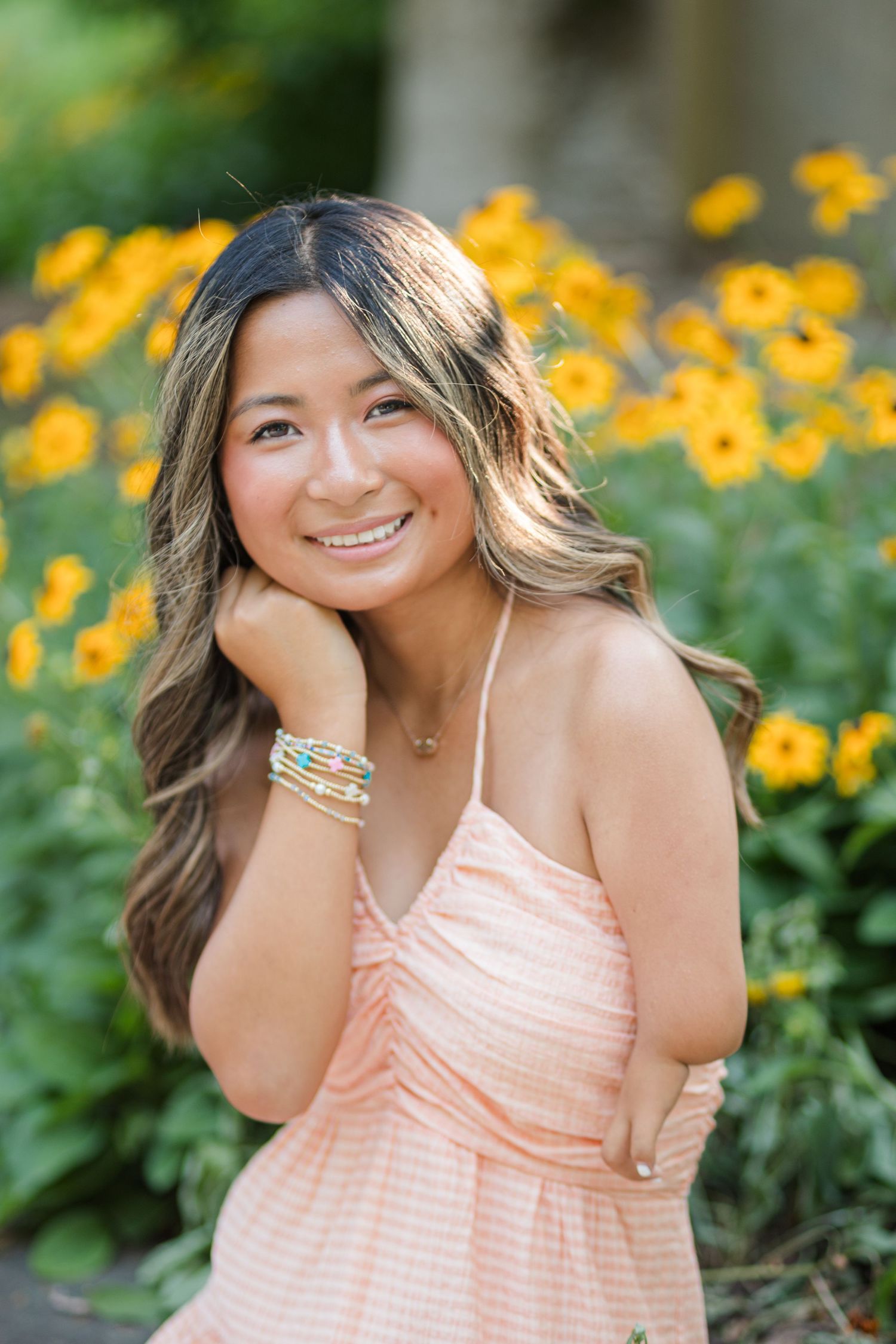 A smiling young woman in a peach dress poses with yellow flowers in a sunny garden background.