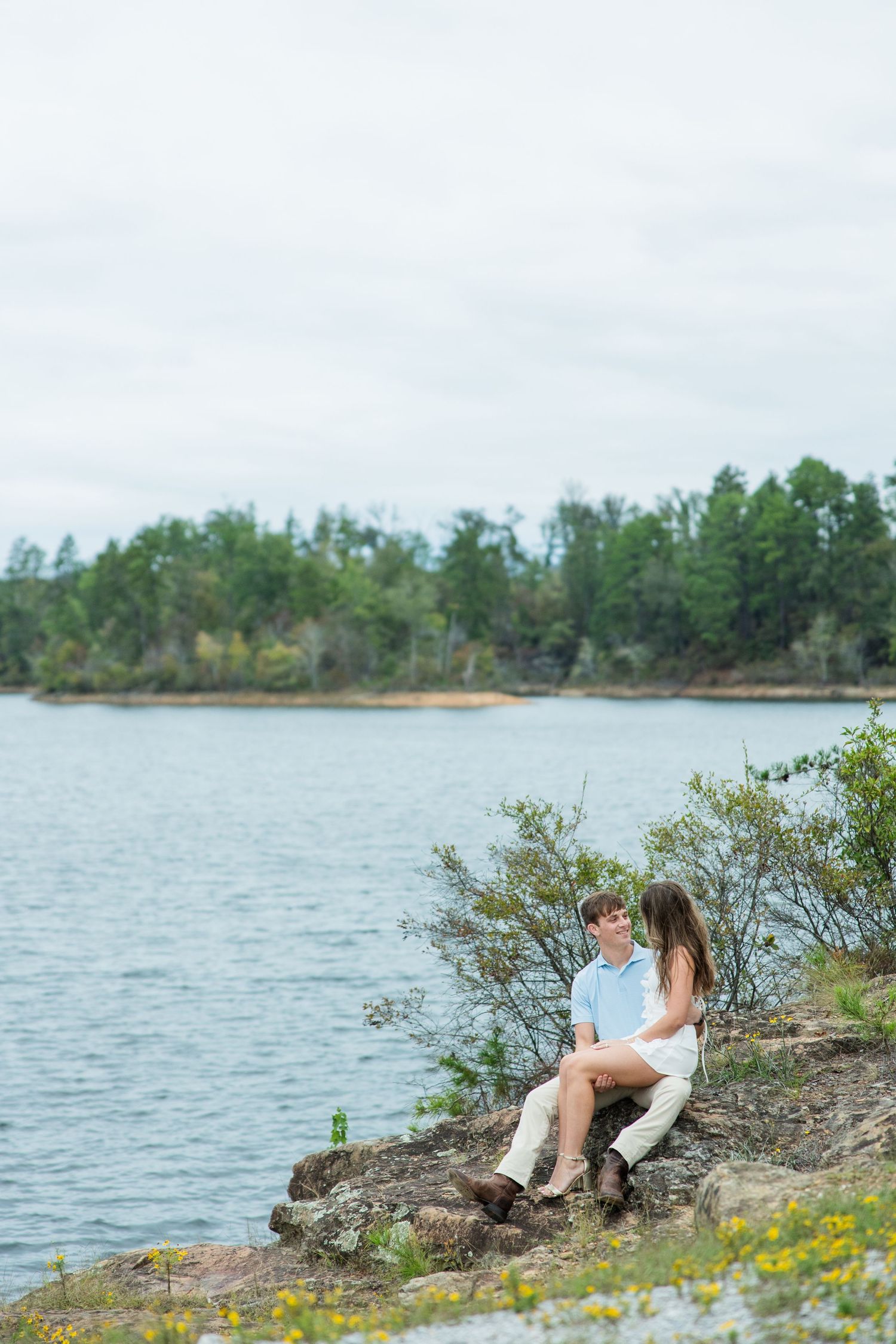 Couple sitting close together on a rocky lakeside outcrop with yellow wildflowers, gazing at each other near calm water and evergreen trees.