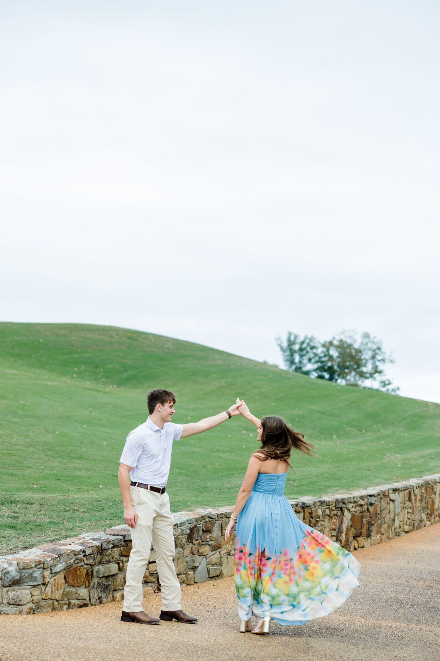 Couple dancing and twirling on a stone-walled path in front of a green grassy hill, she wears a colorful floral skirt.