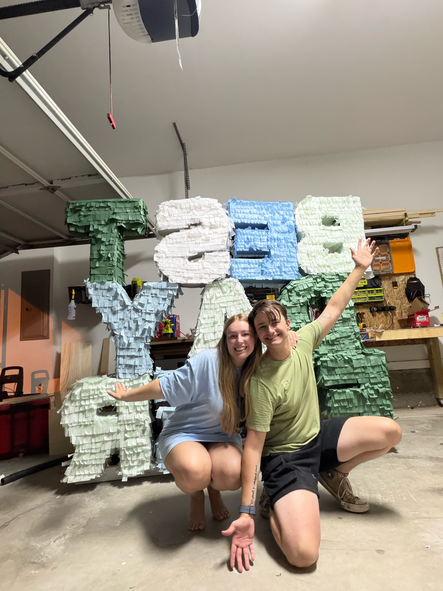 Two women posing in a garage in front of large colorful letter-shaped pinatas spelling a name.