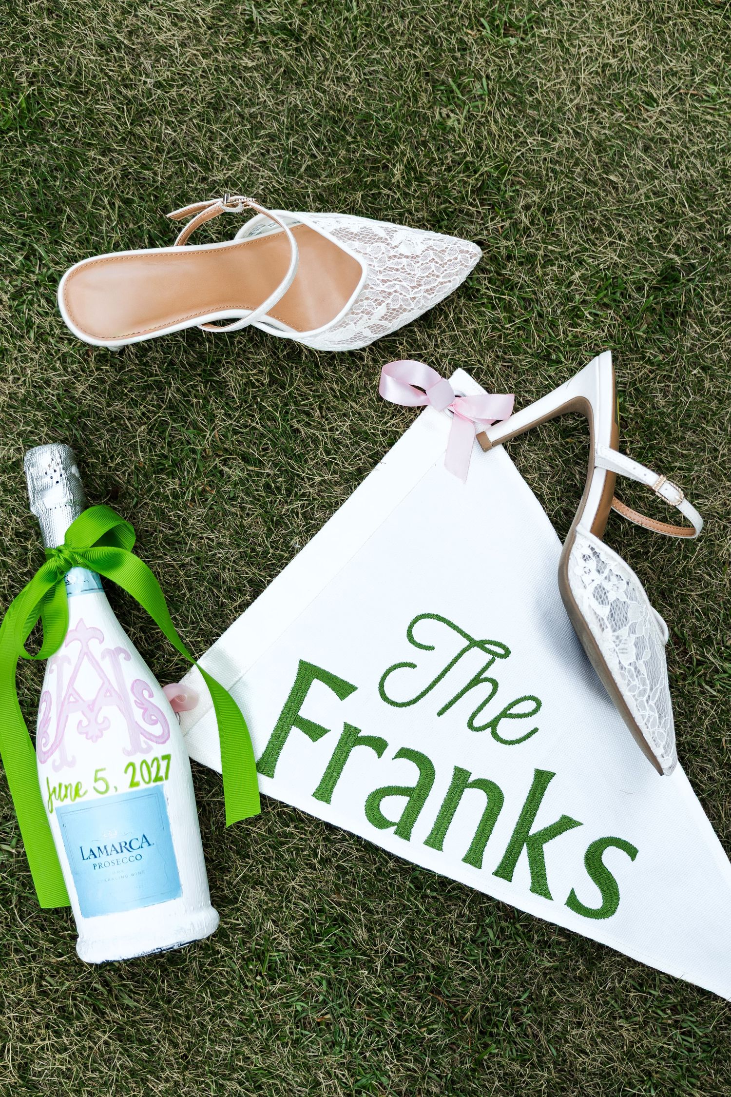 Wedding flat lay with lace heels, champagne bottle with green ribbon, and The Franks pennant flag on green grass.