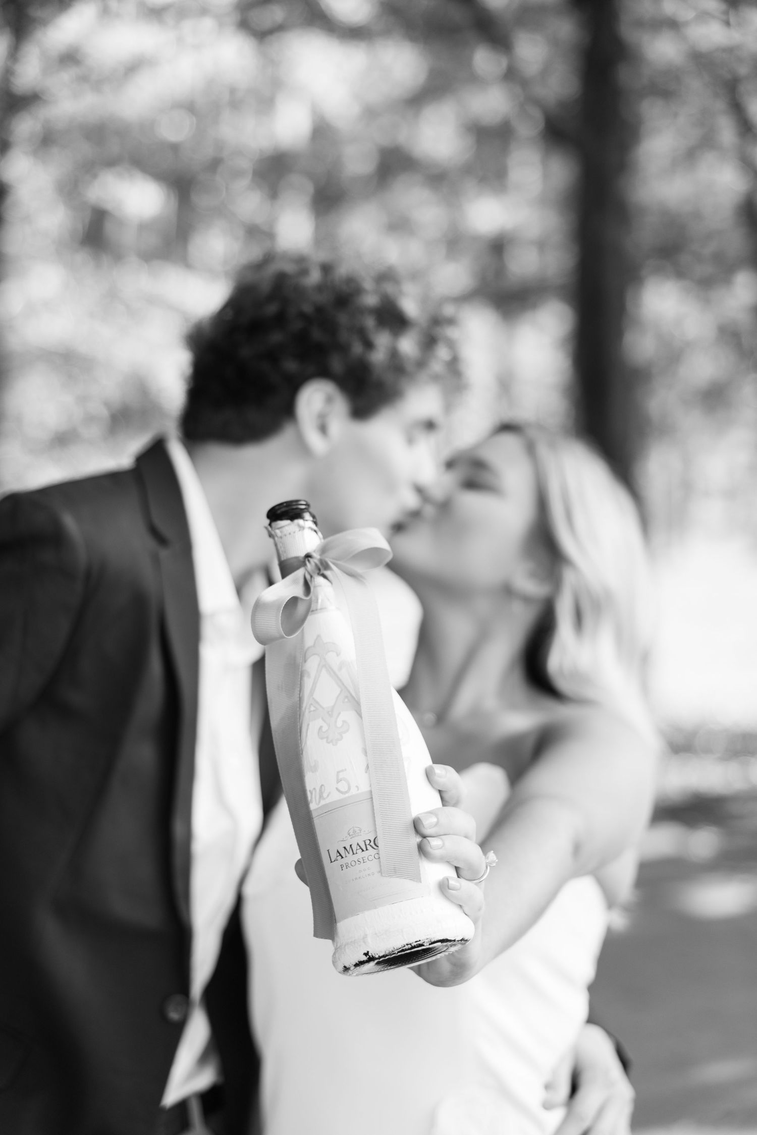 Black and white photo of newlyweds kissing behind a Lanaire champagne bottle outdoors.