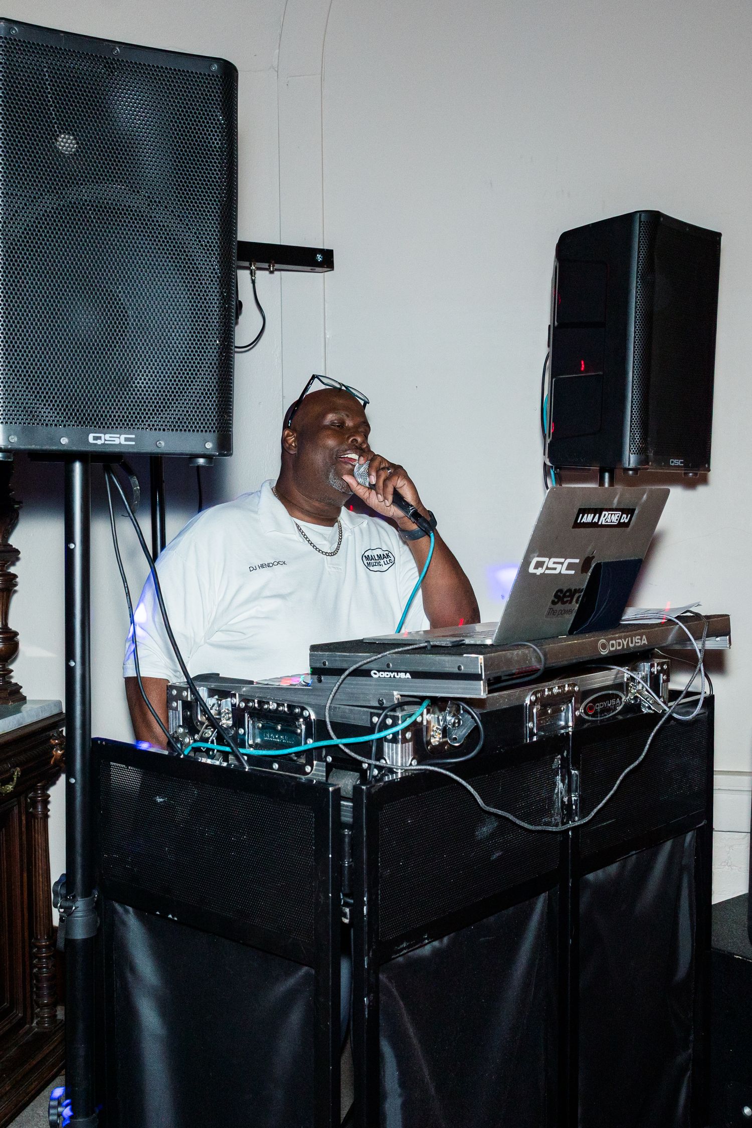 DJ performing at wedding reception behind equipment setup with QSC speakers and mixing console.