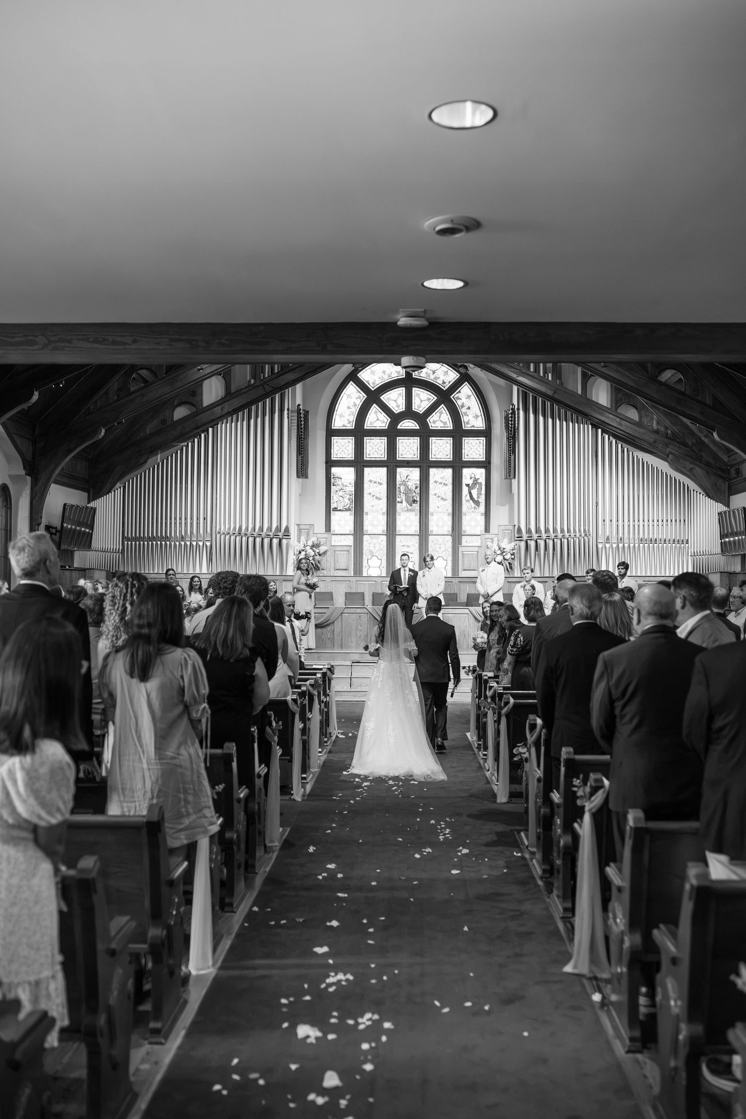 Black and white wide-angle view of a wedding ceremony inside a traditional church with guests seated in wooden pews.