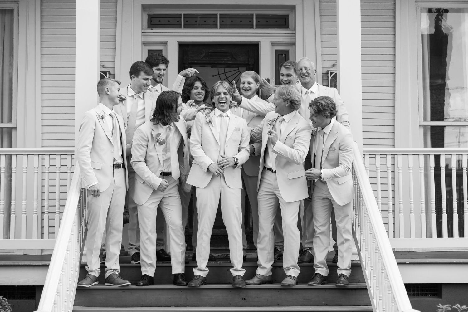 Black and white photo of a wedding party in light suits posing on porch steps of a classic white house.