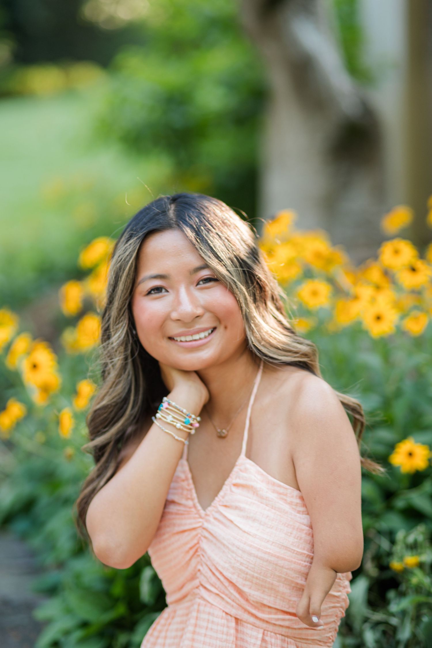 Smiling young woman in peach dress with bracelets posing near yellow flowers outdoors.