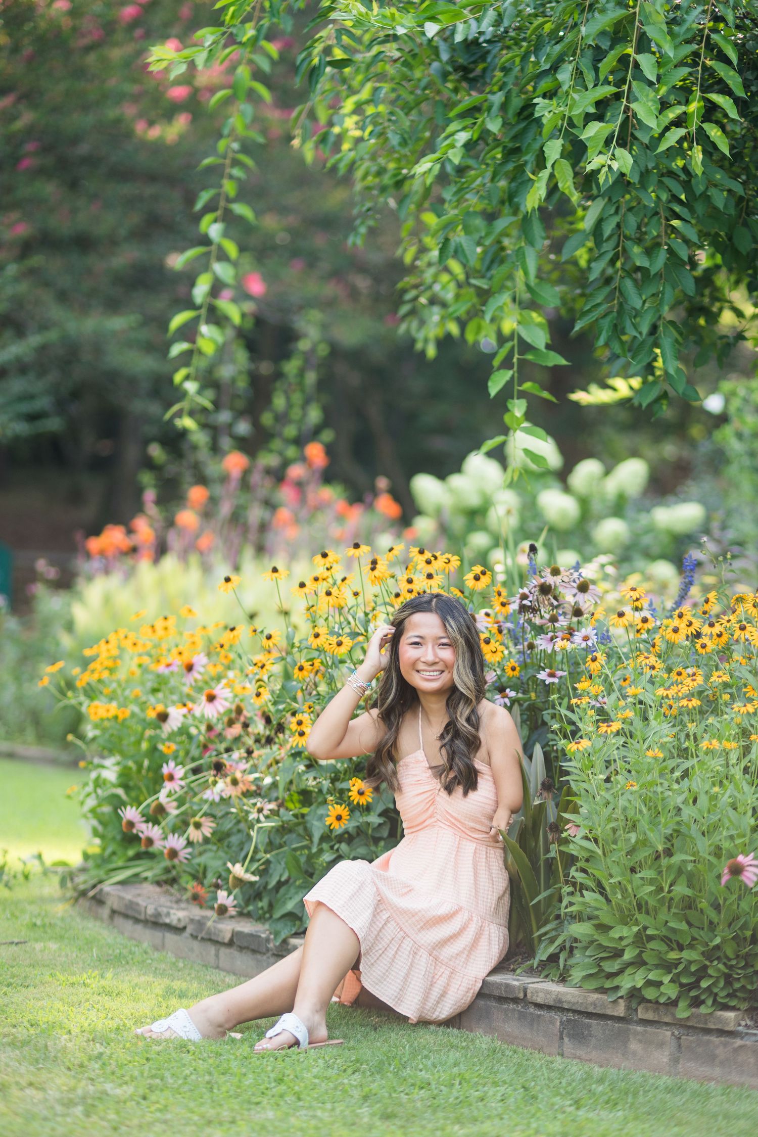 Young woman in pink dress sitting on garden edge surrounded by colorful blooming flowers and lush green trees.