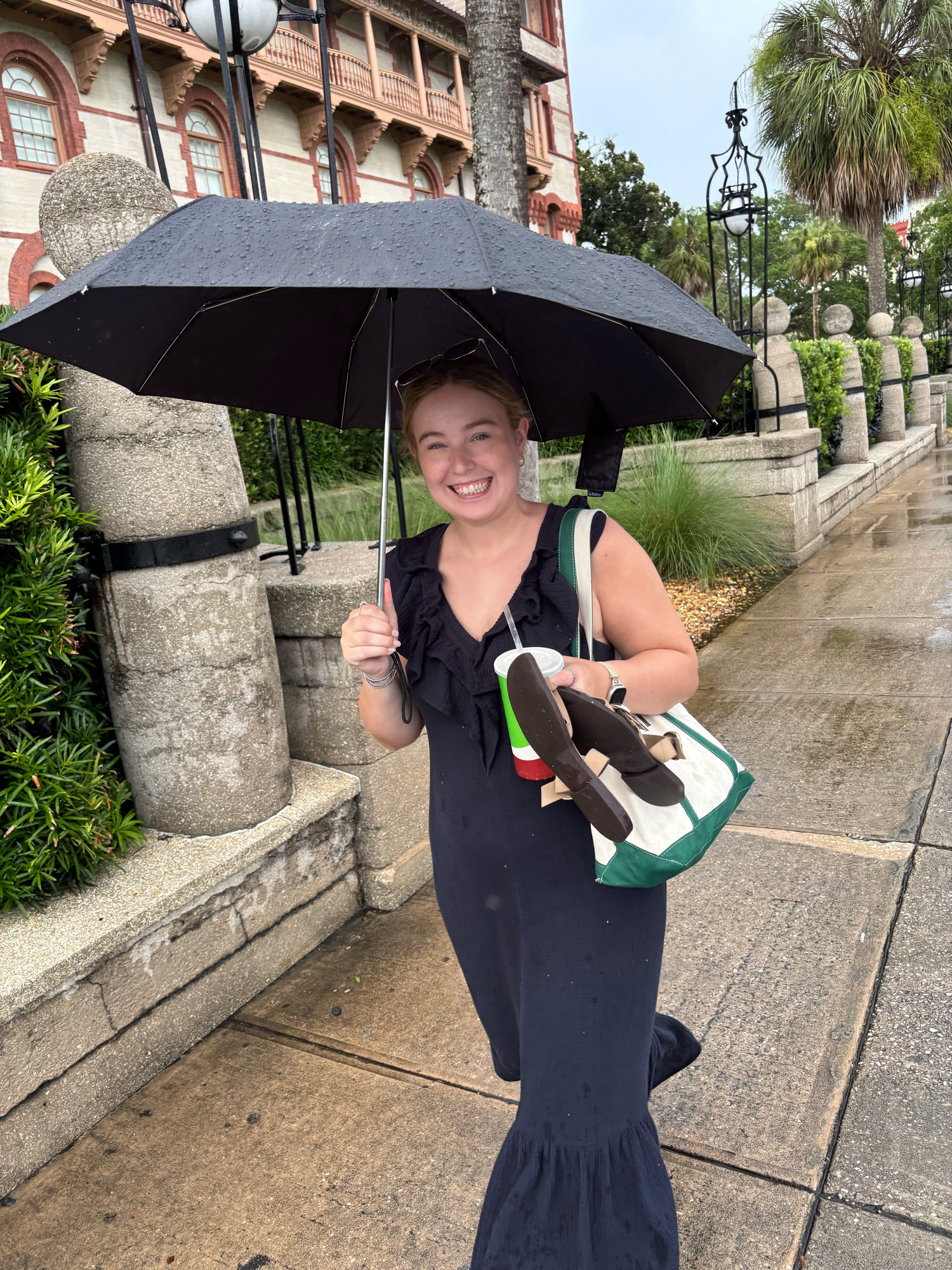 Happy woman with black umbrella and colorful tote bag smiling on rainy historic sidewalk.