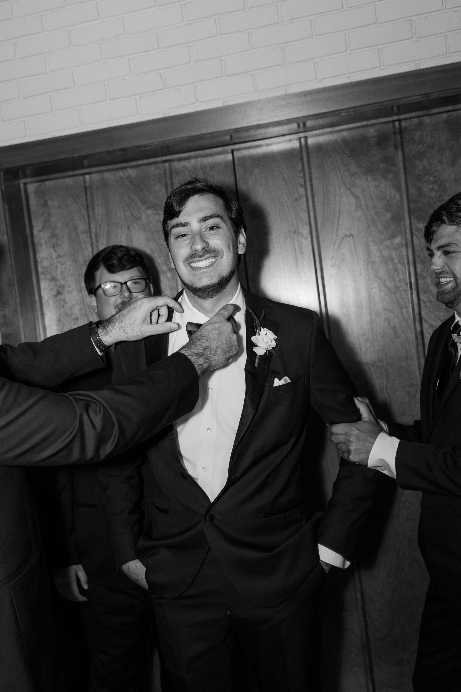 Black and white photo of a smiling groom in a tuxedo being helped with his bow tie by groomsmen.