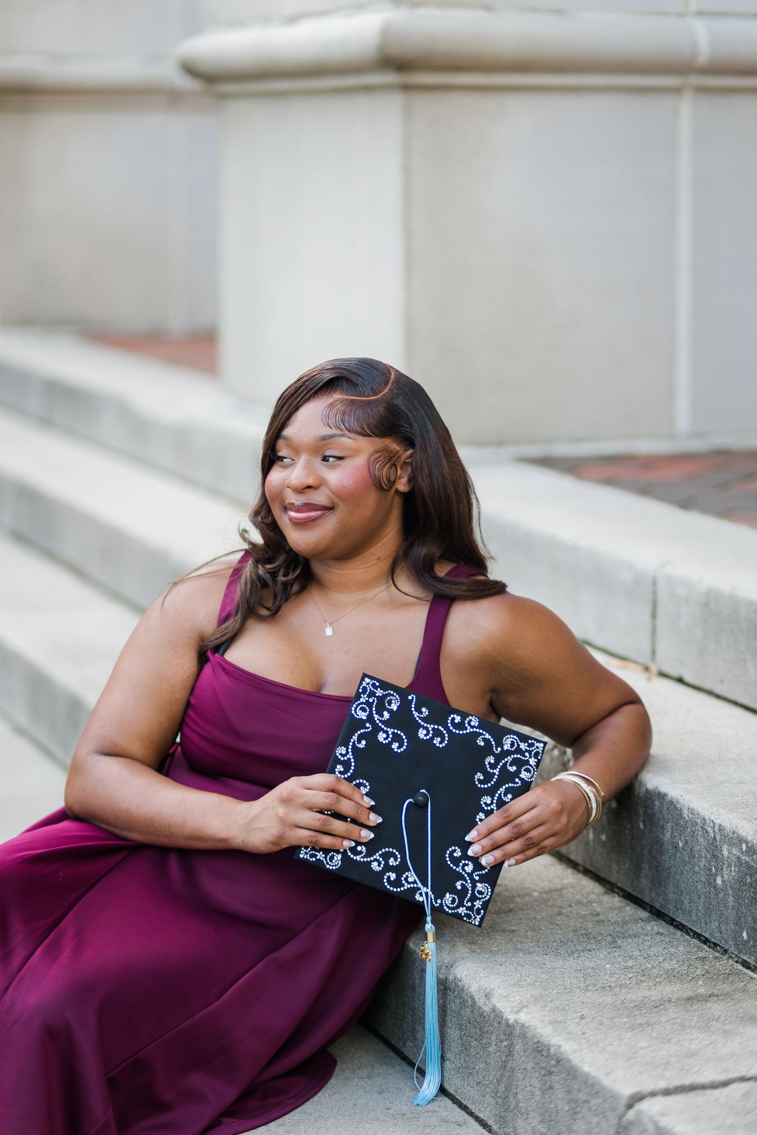 Young woman in burgundy gown holding decorated graduation cap while sitting on stone steps.
