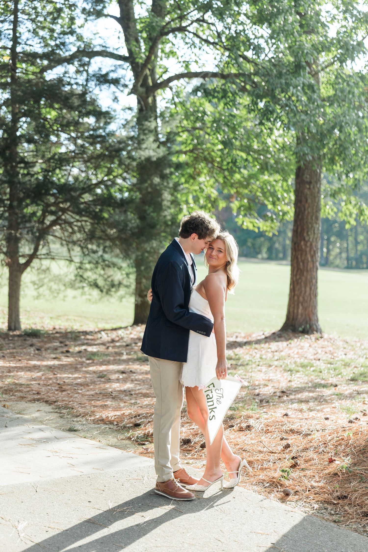 Couple embracing on a shaded path near a golf course, bride in white dress holding an engagement pennant.