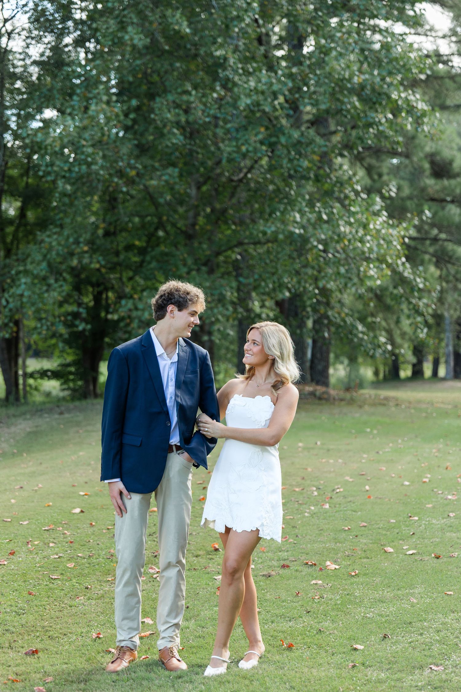 Engaged couple standing together on a green golf course, man in navy blazer, woman in white mini dress.