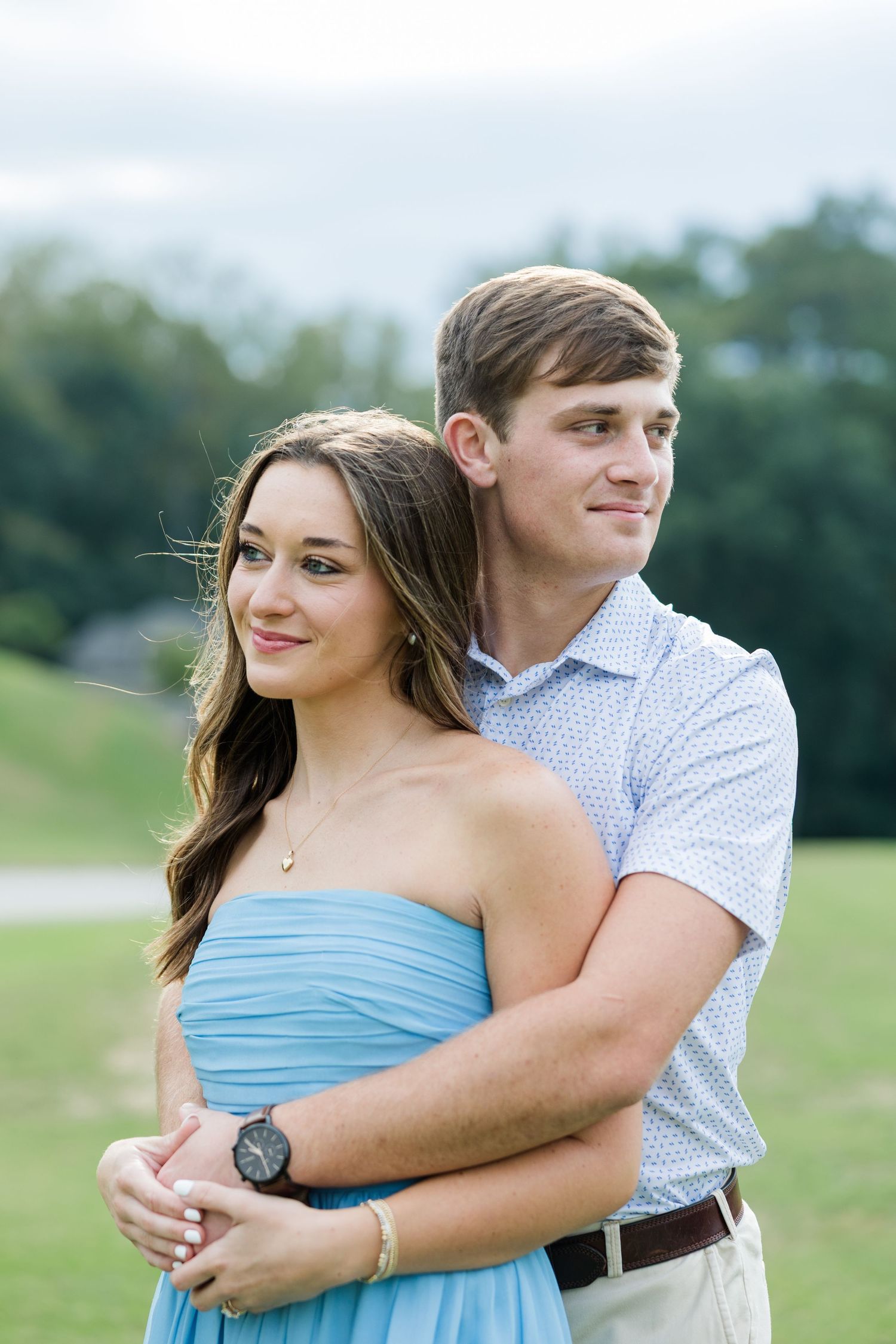 Smiling couple embracing outdoors in a grassy park, the woman in a blue strapless dress and the man in a patterned polo shirt.
