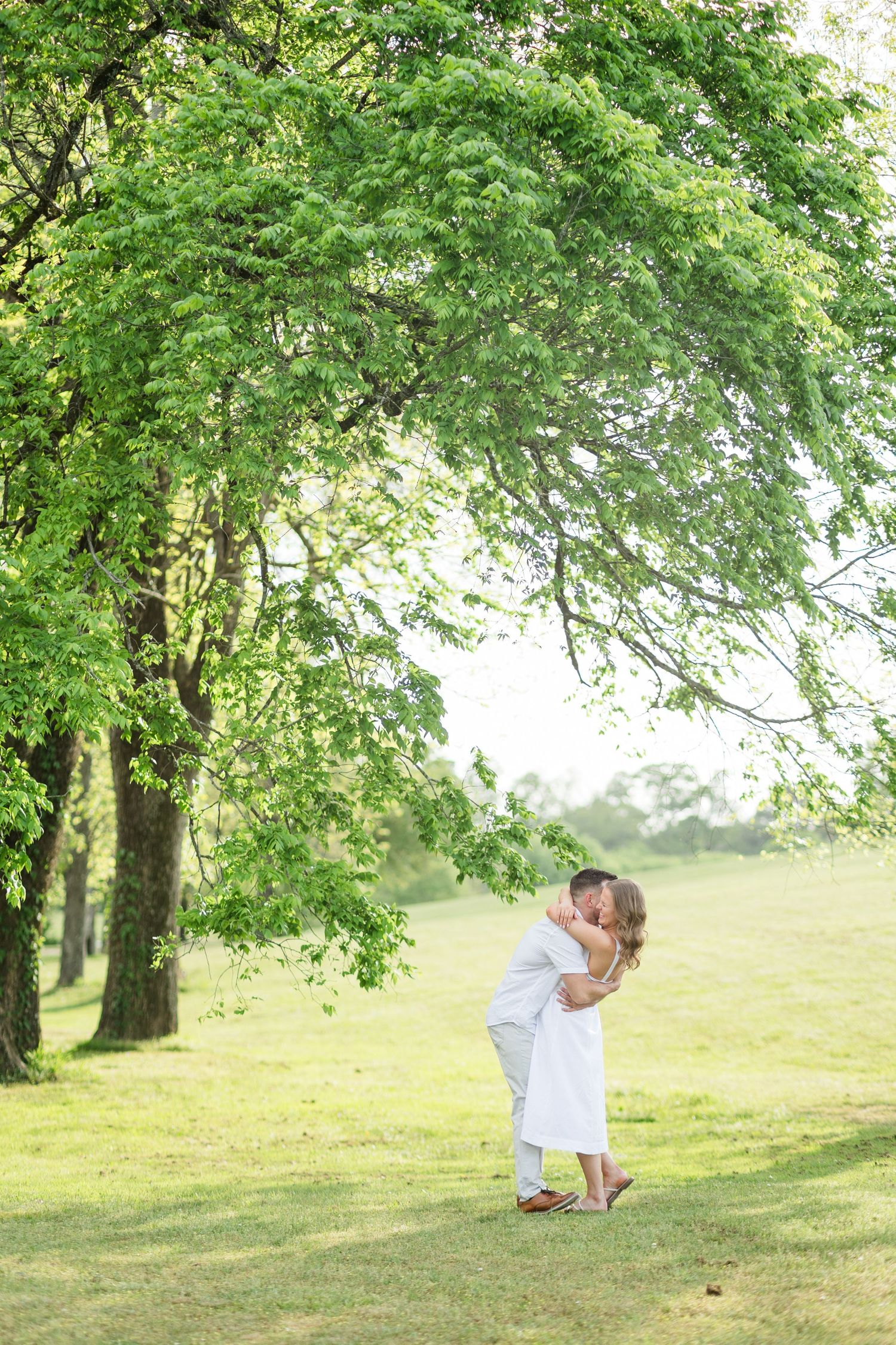 Couple sharing a kiss under lush green trees in a sunlit park during engagement shoot.