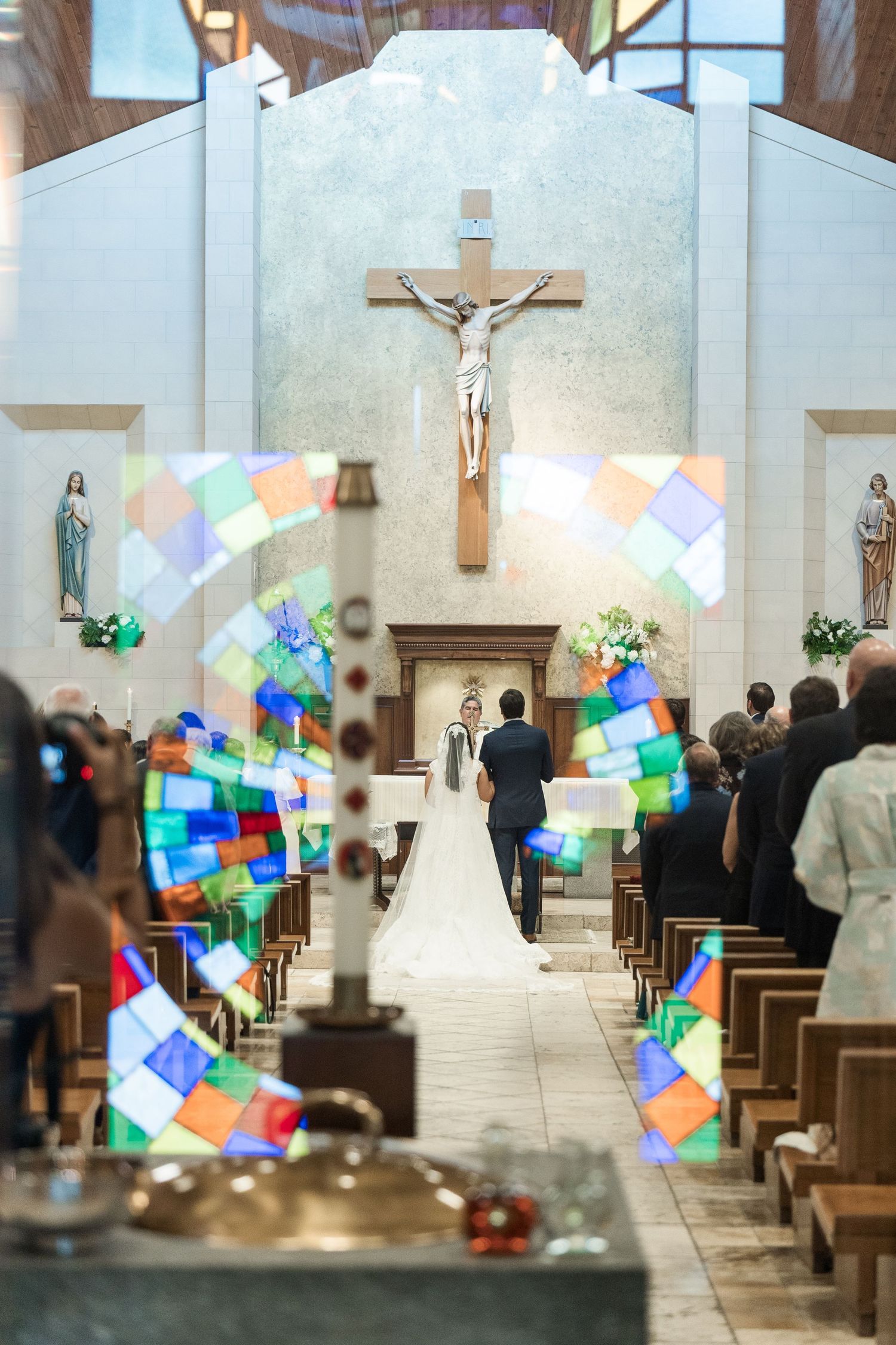 A bride and groom walk down the aisle of a Catholic church adorned with colorful stained glass and a crucifix.