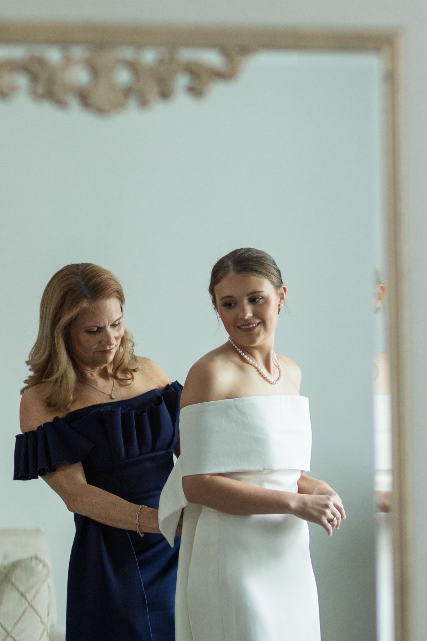 A bride in a white off-shoulder gown being helped by a woman in a navy dress in an elegant room.