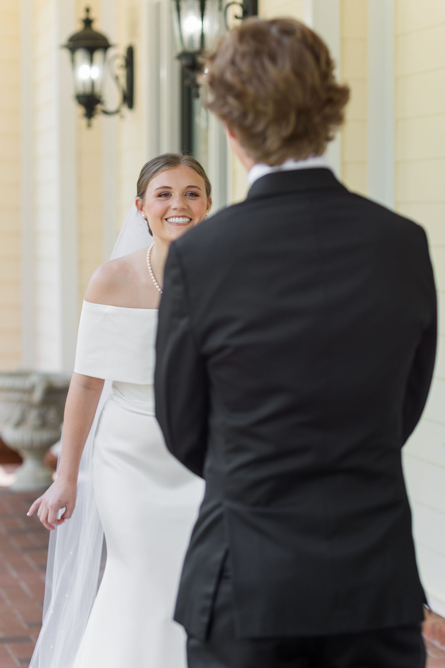 Smiling bride in white off-shoulder gown with veil faces groom in black tuxedo outdoors.