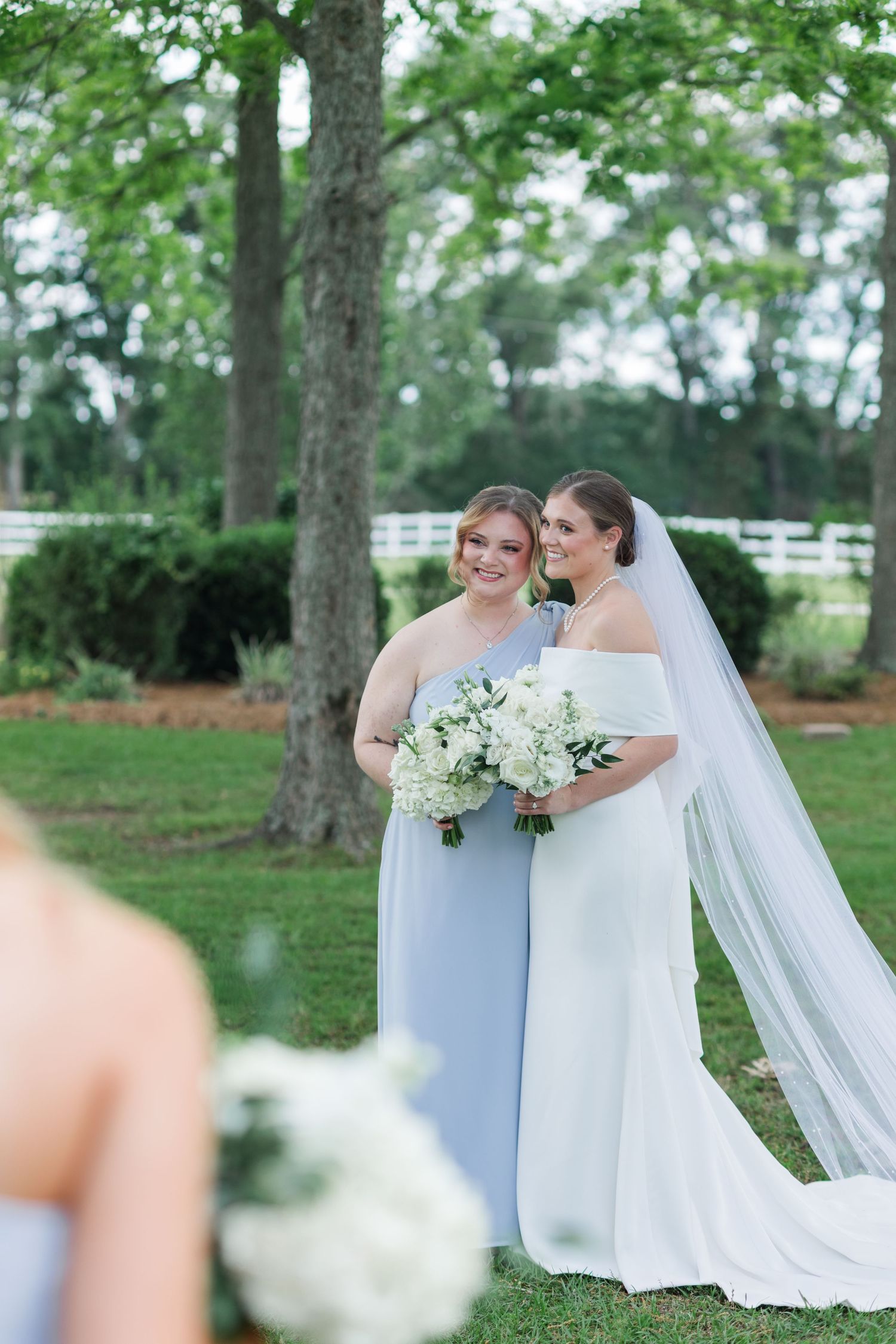 Bride in white off-shoulder gown with veil stands beside bridesmaid in blue dress holding floral bouquets outdoors.
