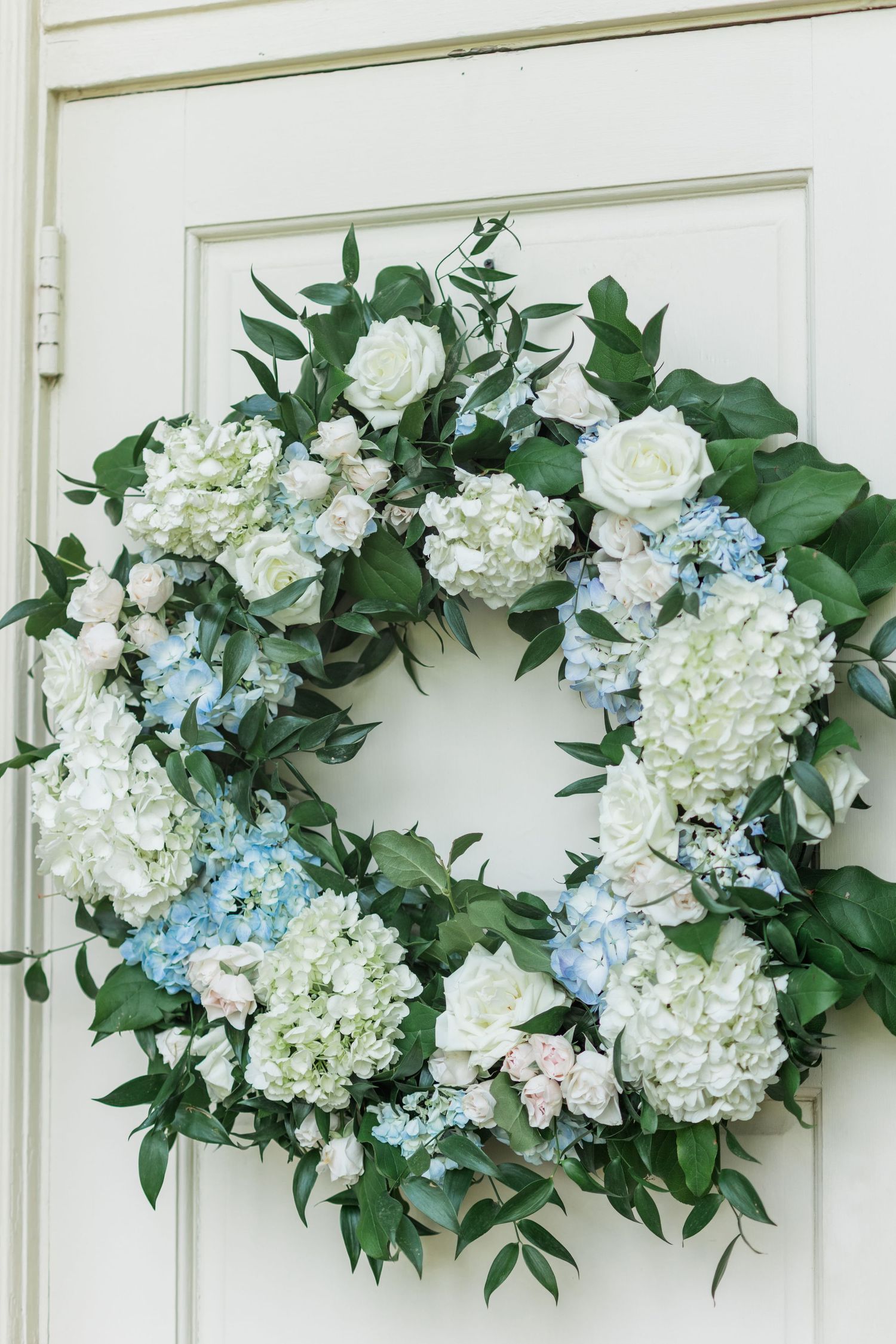 Elegant wedding wreath with white roses, blue and white hydrangeas, and greenery hanging on a white door.