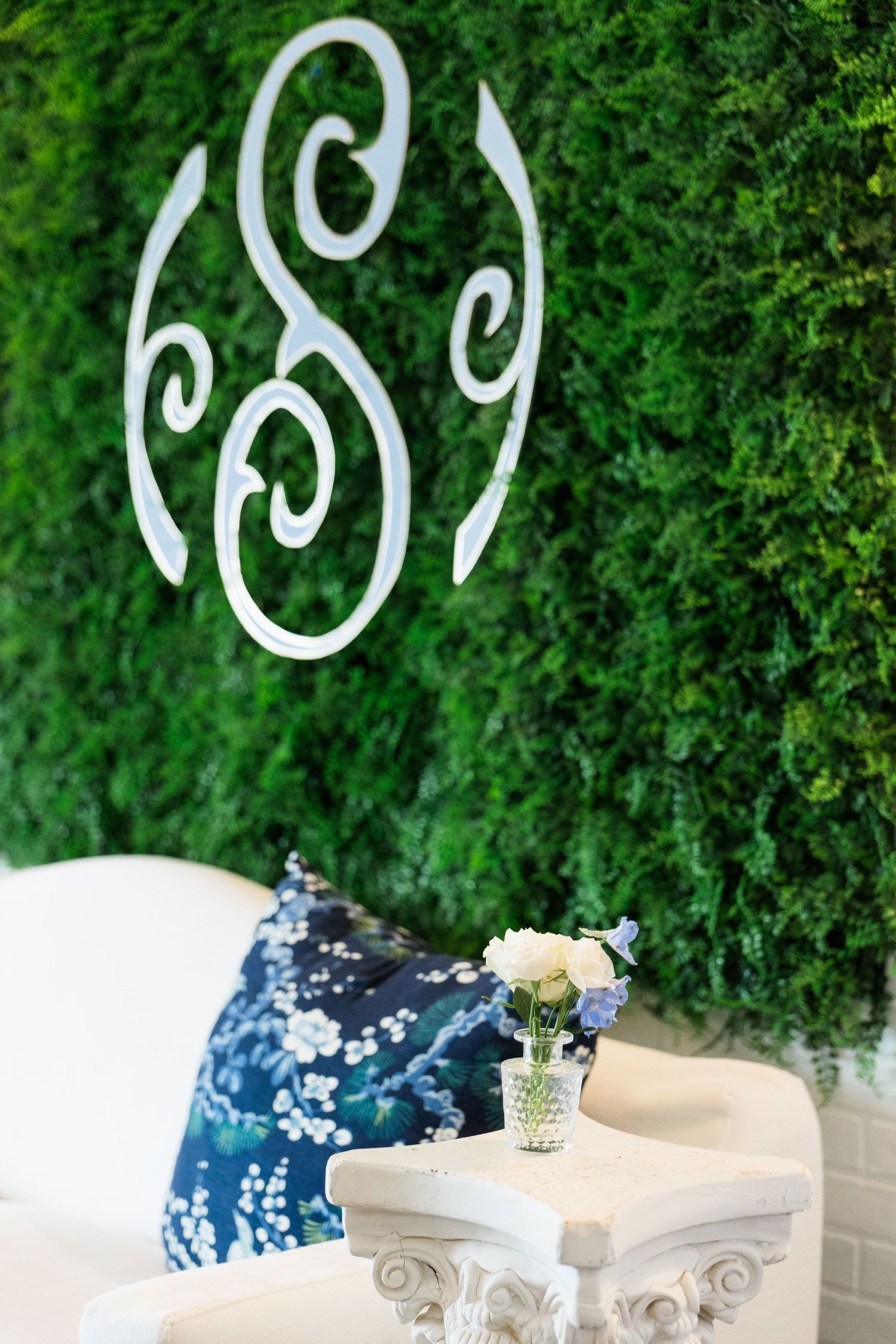 White monogram letters displayed on lush green hedge wall above elegant white lounge seating.