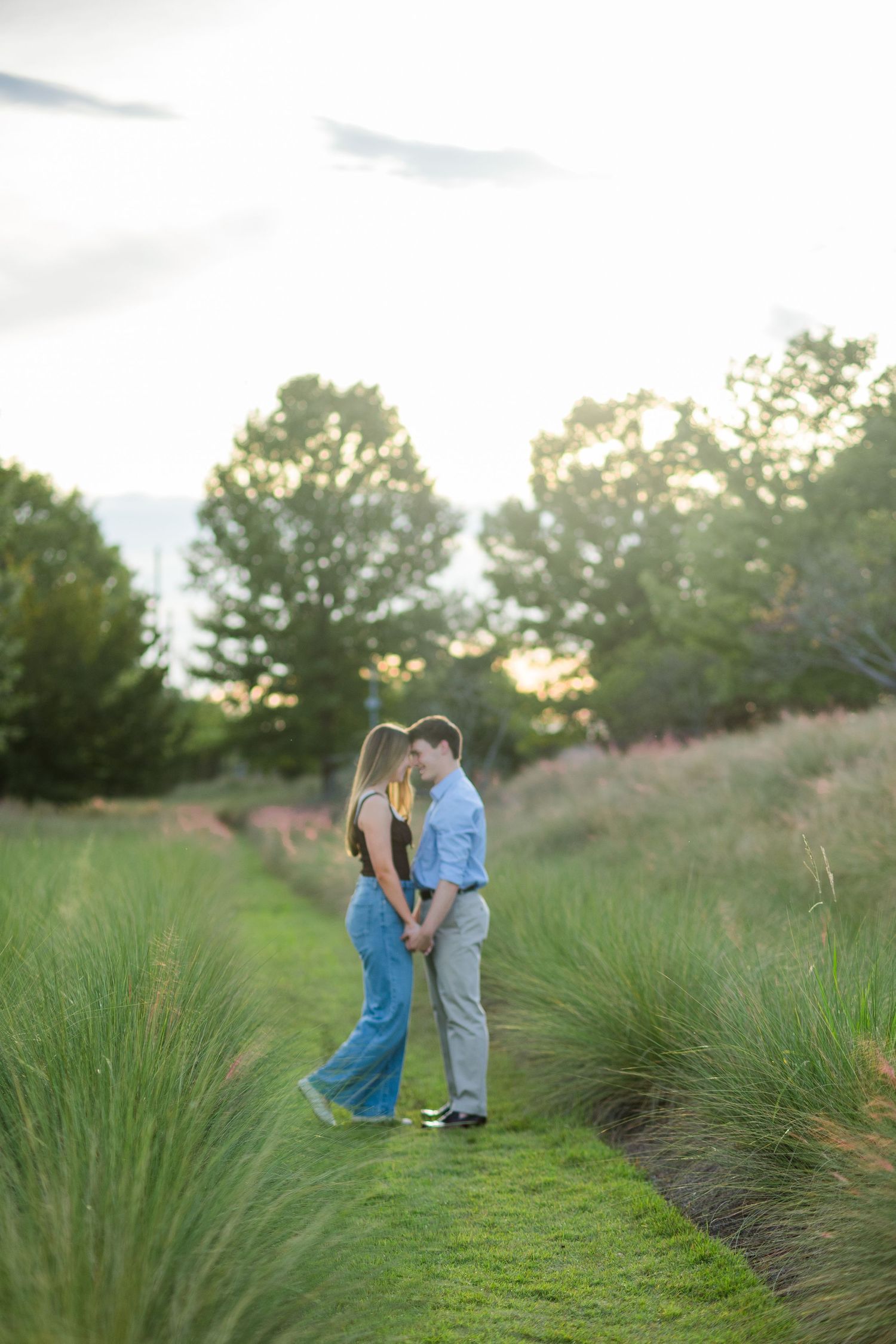 A couple stands face-to-face on a grassy path surrounded by tall ornamental grasses at golden hour.
