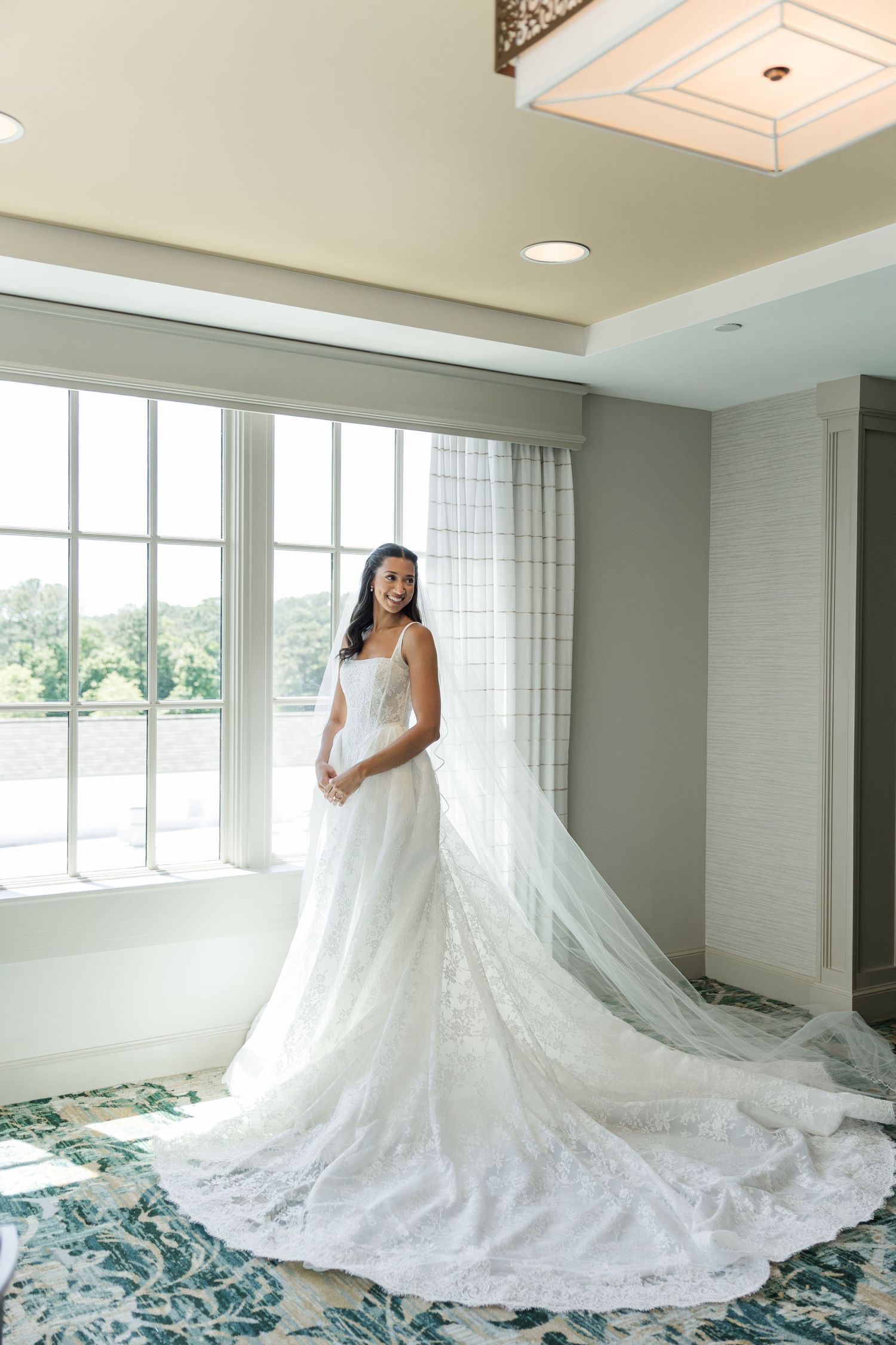 Bride in a white lace wedding gown with long veil stands by large windows in an elegant hotel suite.