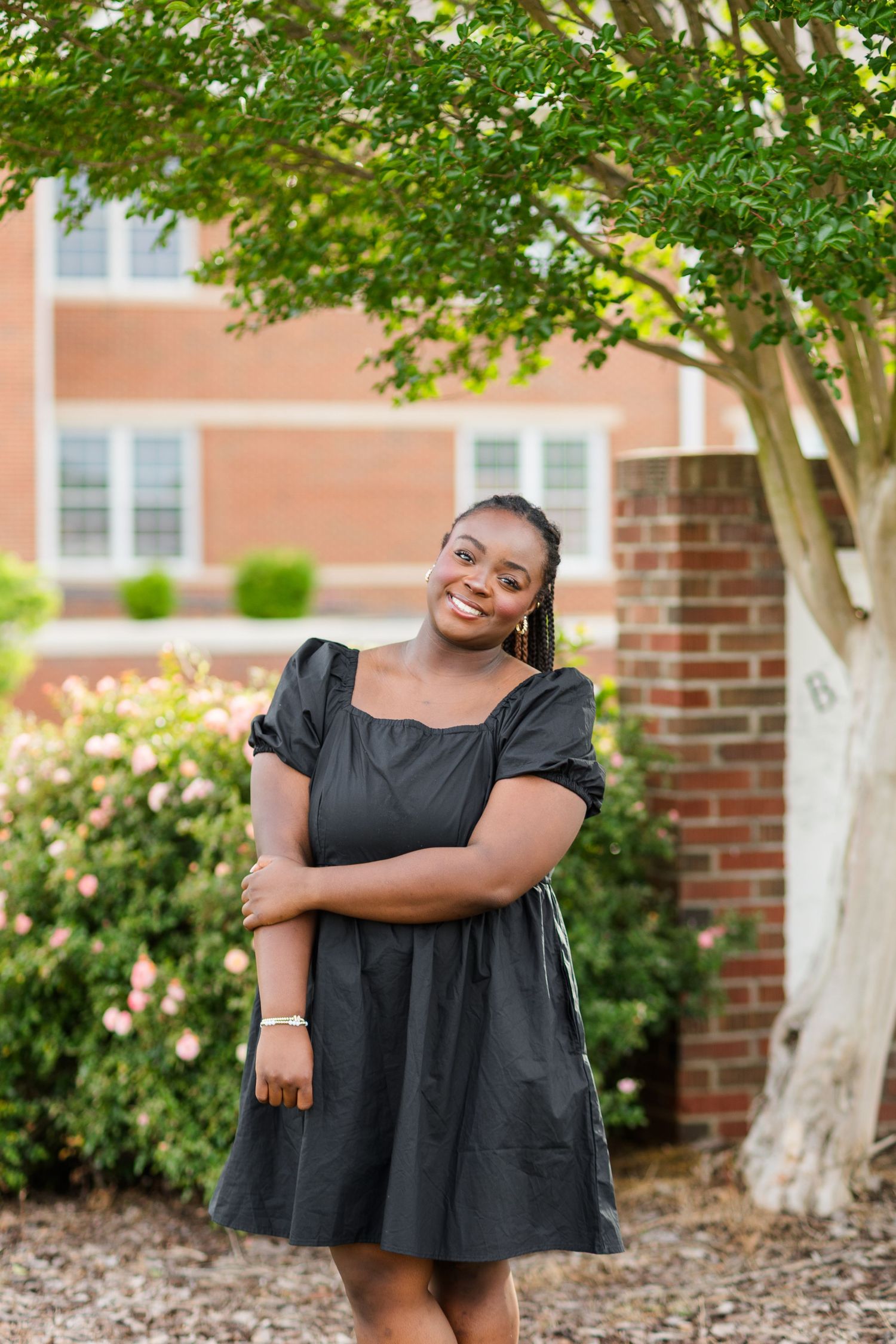 Young Black woman in black dress smiling outdoors near brick building and flowering bushes.