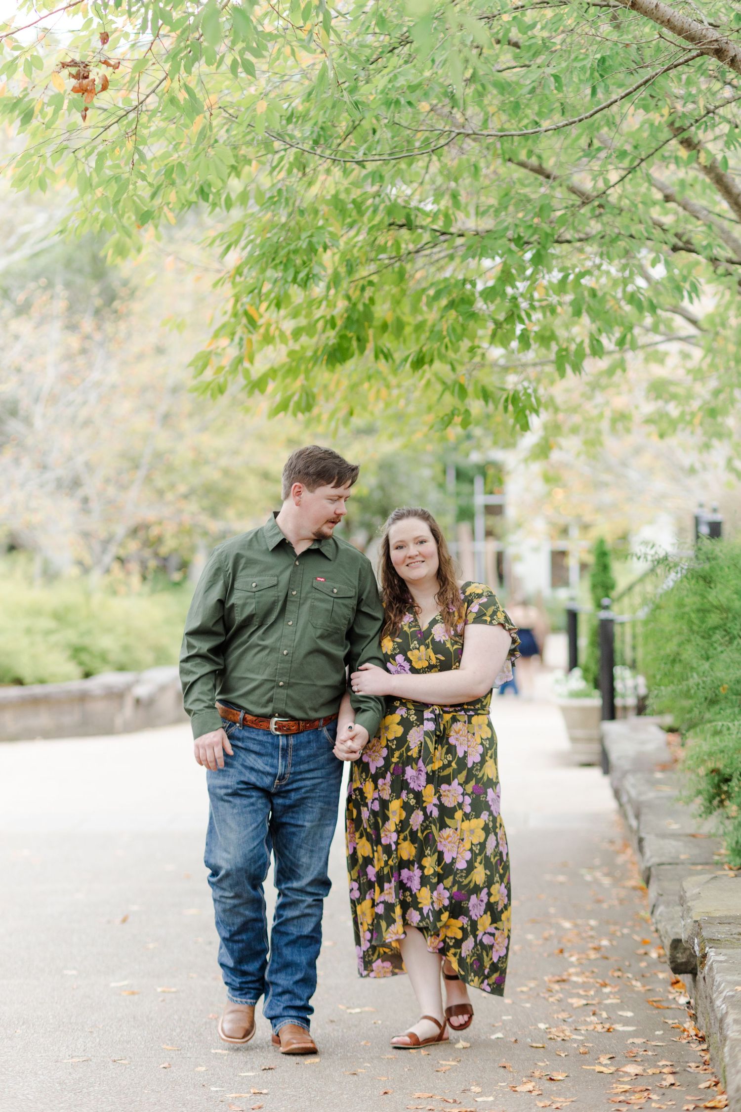 A couple walks hand-in-hand along a tree-lined path, smiling at each other in autumn surroundings.