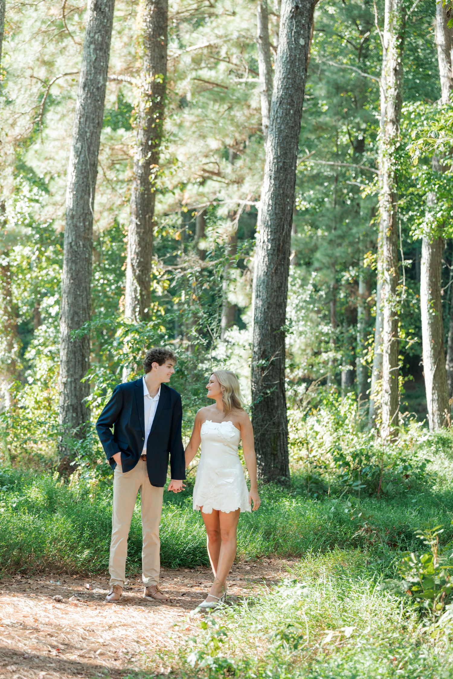 Couple holding hands walking through sunlit pine forest, woman in white dress and man in navy blazer.