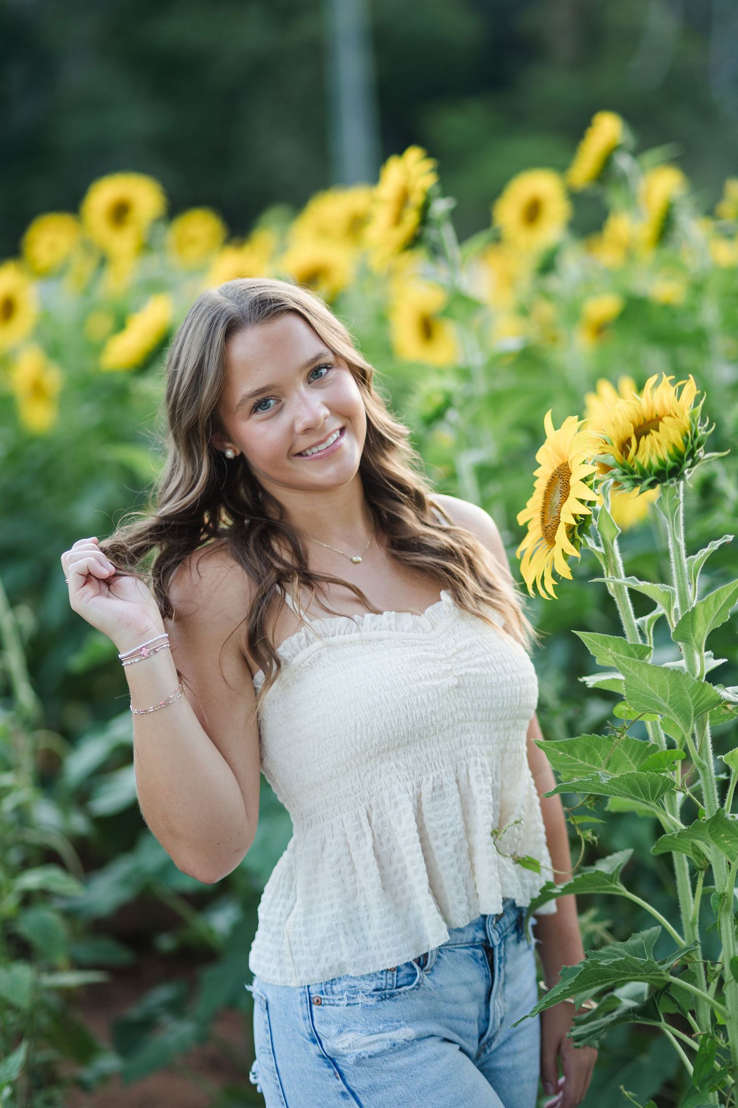 Smiling young woman in white top and jeans posing in a vibrant sunflower field on a sunny day.