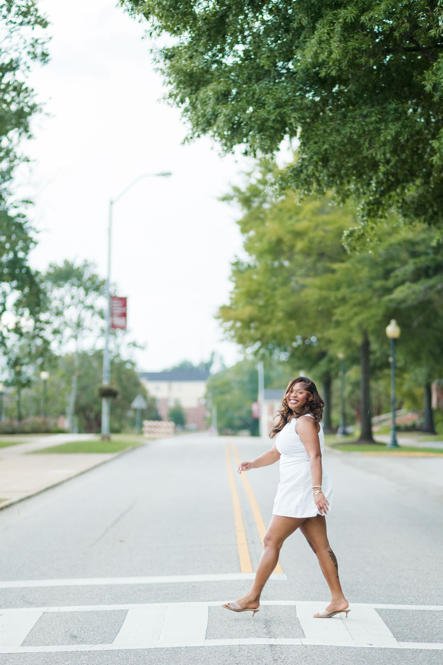 A young woman in a white mini dress walks across a crosswalk on a tree-lined street, smiling over her shoulder.