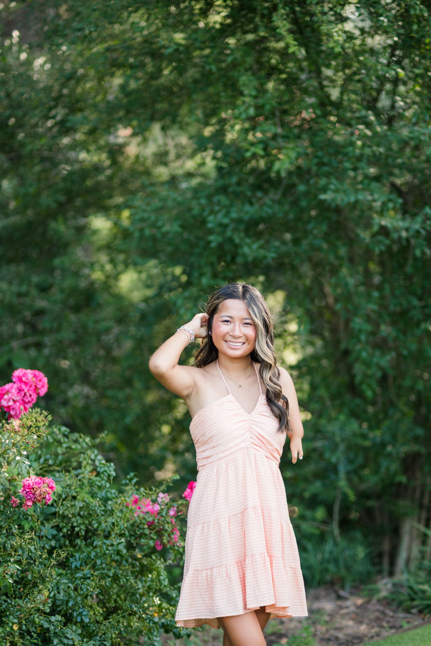 Smiling young woman in pink dress posing outdoors near blooming roses with lush green trees in background.