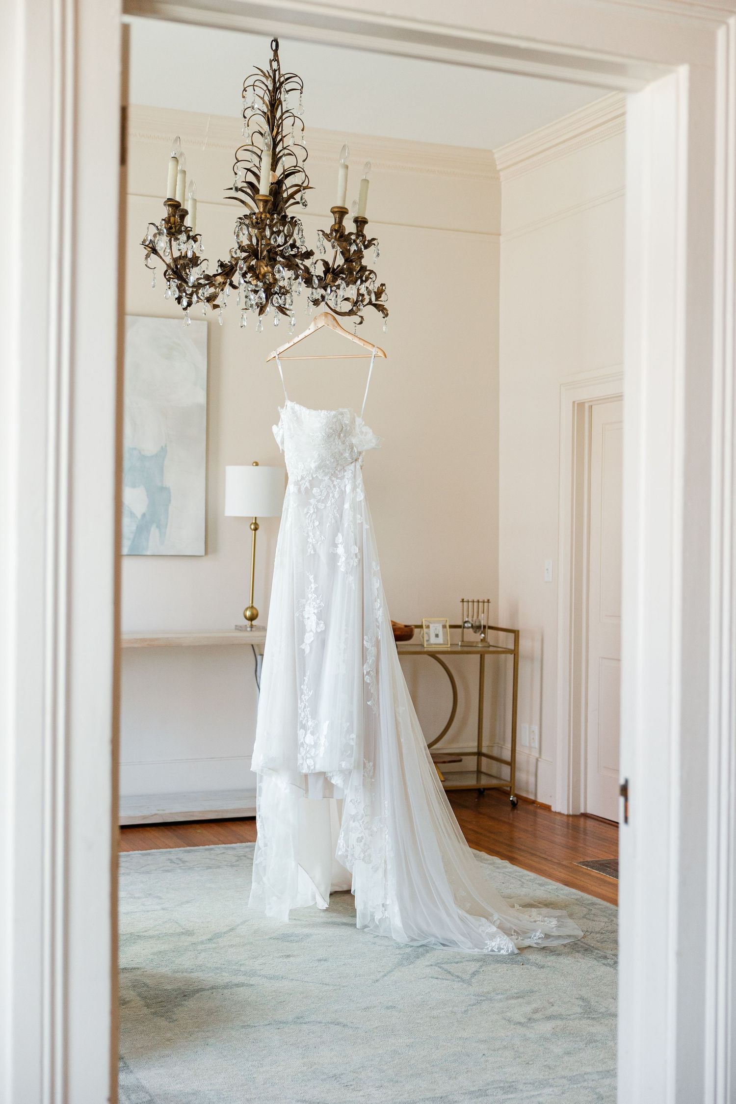 A white lace wedding dress with a long train hanging from a chandelier in an elegant, softly lit bridal suite.