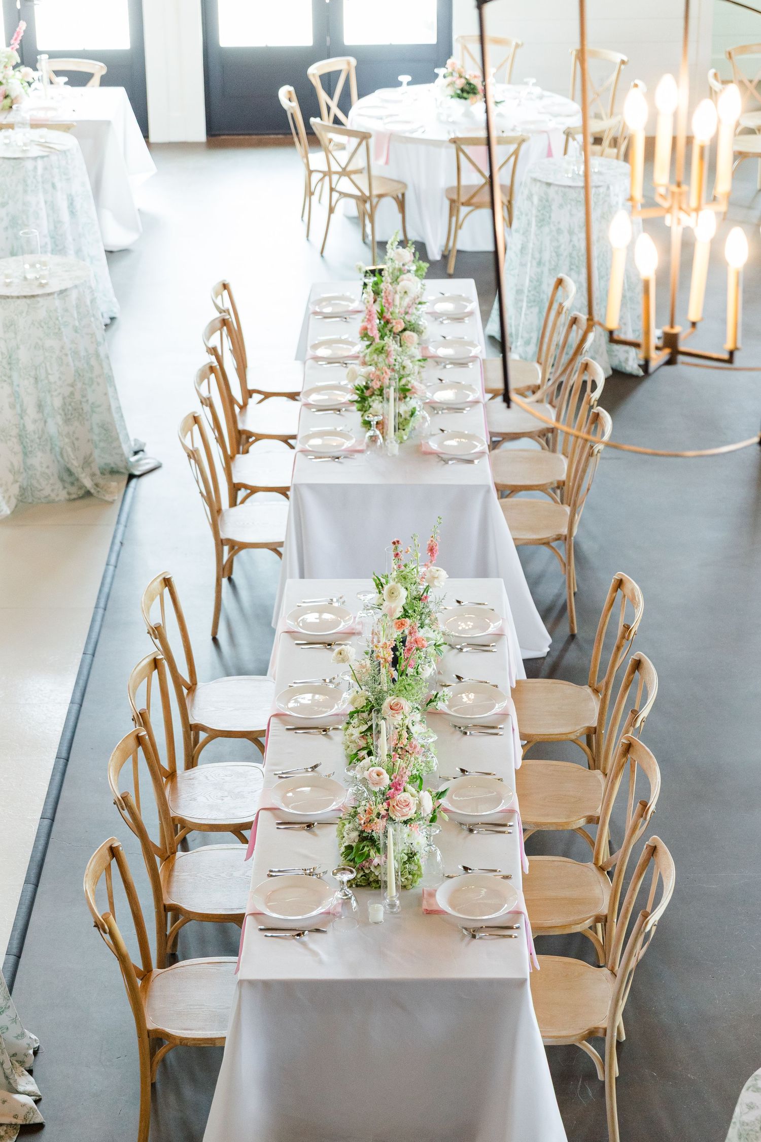 Elegant wedding reception table with floral runner, white linens, gold cross-back chairs, and candelabras in a bright venue.