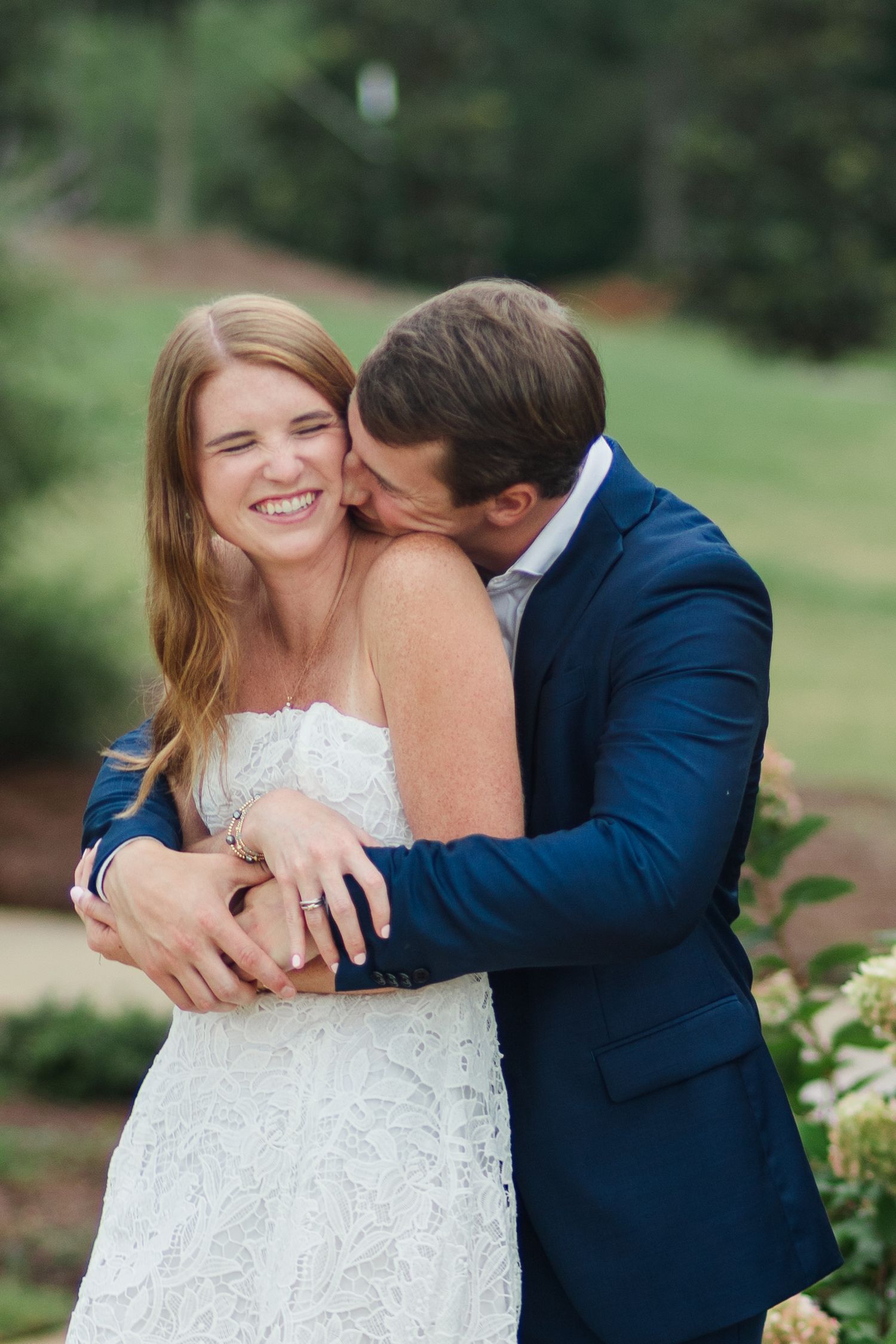 Groom in navy suit kissing bride in lace dress, who smiles joyfully, in an outdoor wedding portrait.