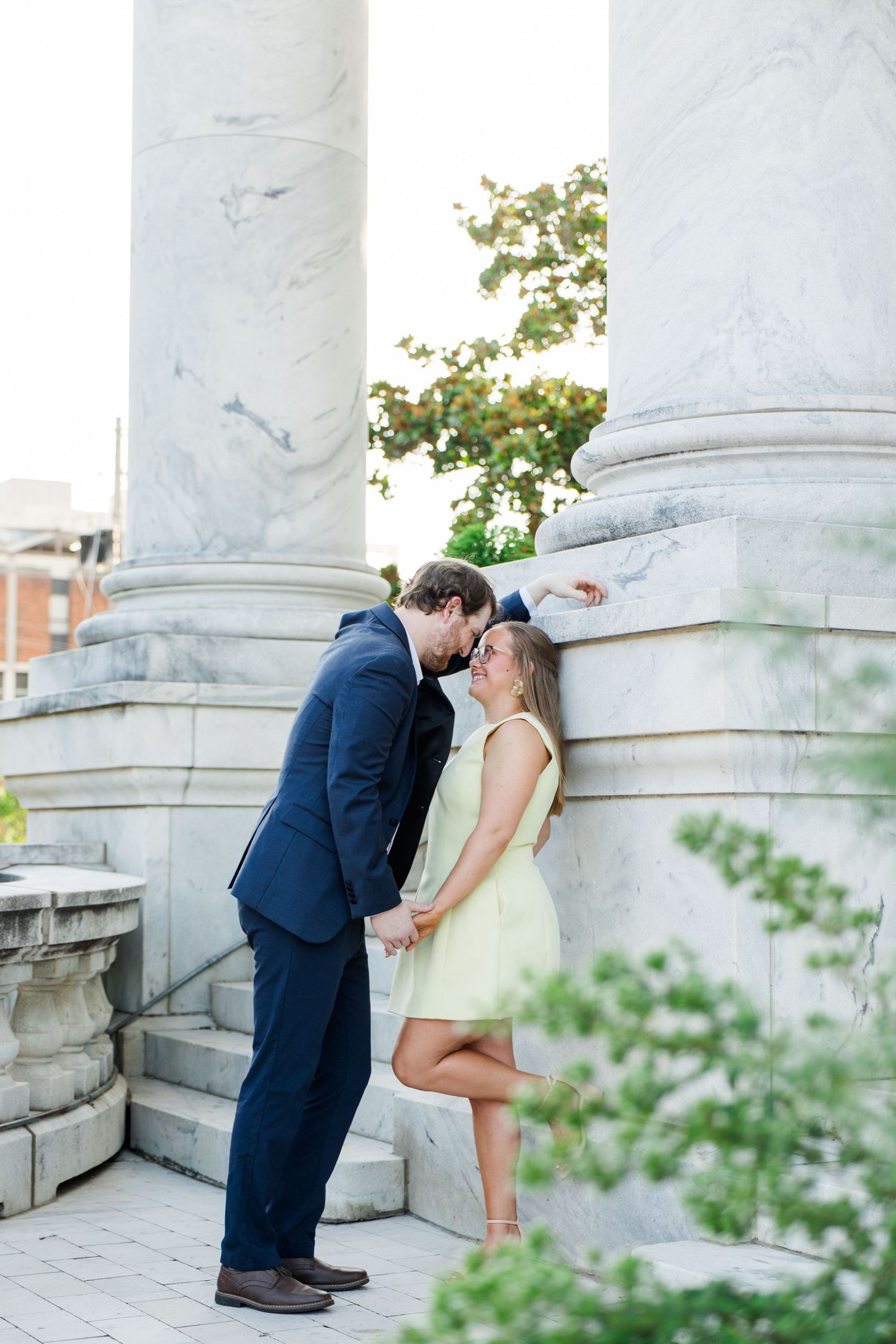 Couple sharing a romantic moment between marble columns, man in navy suit, woman in light green dress.