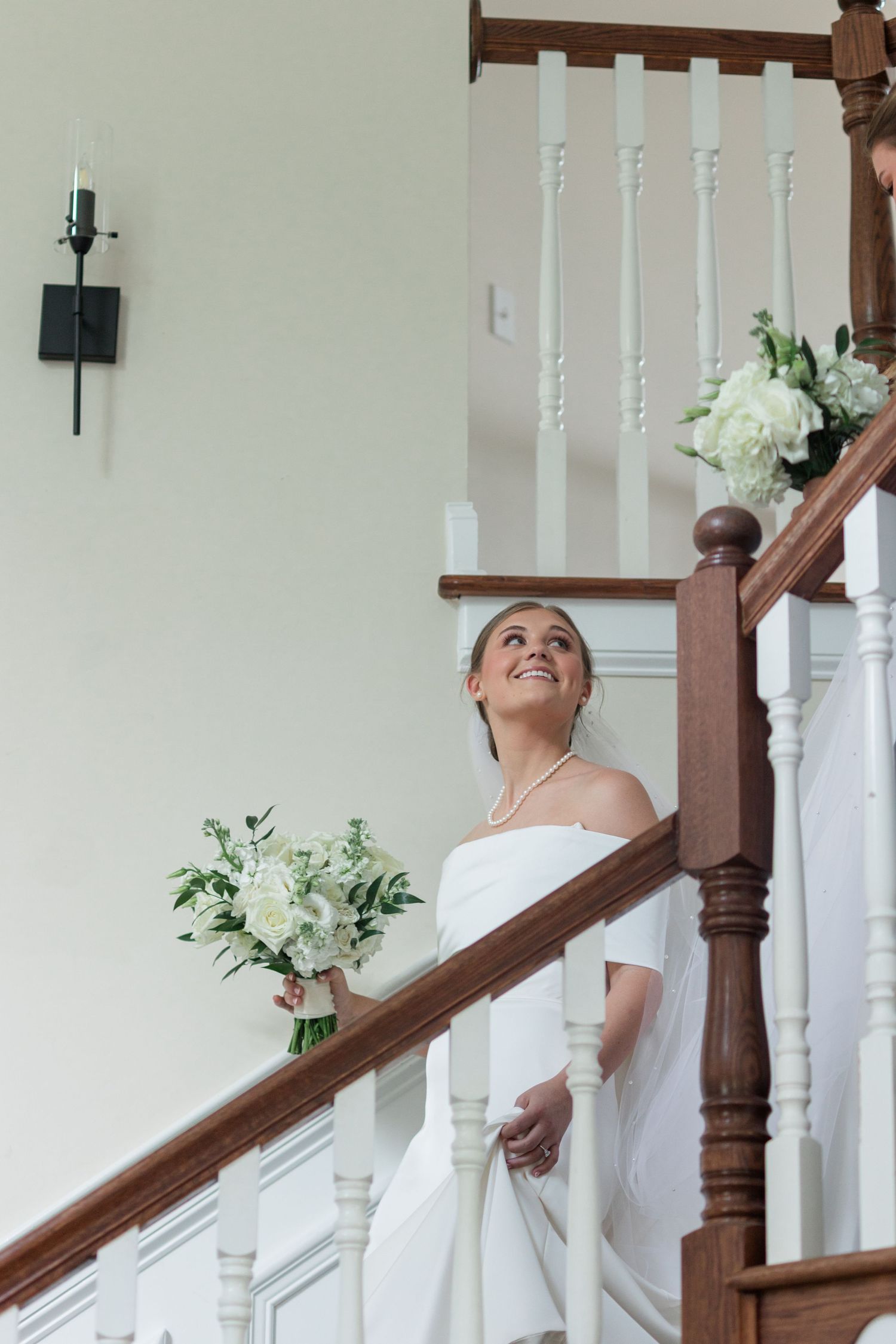 Smiling bride in off-shoulder white gown holds floral bouquet while descending a classic wooden staircase indoors.