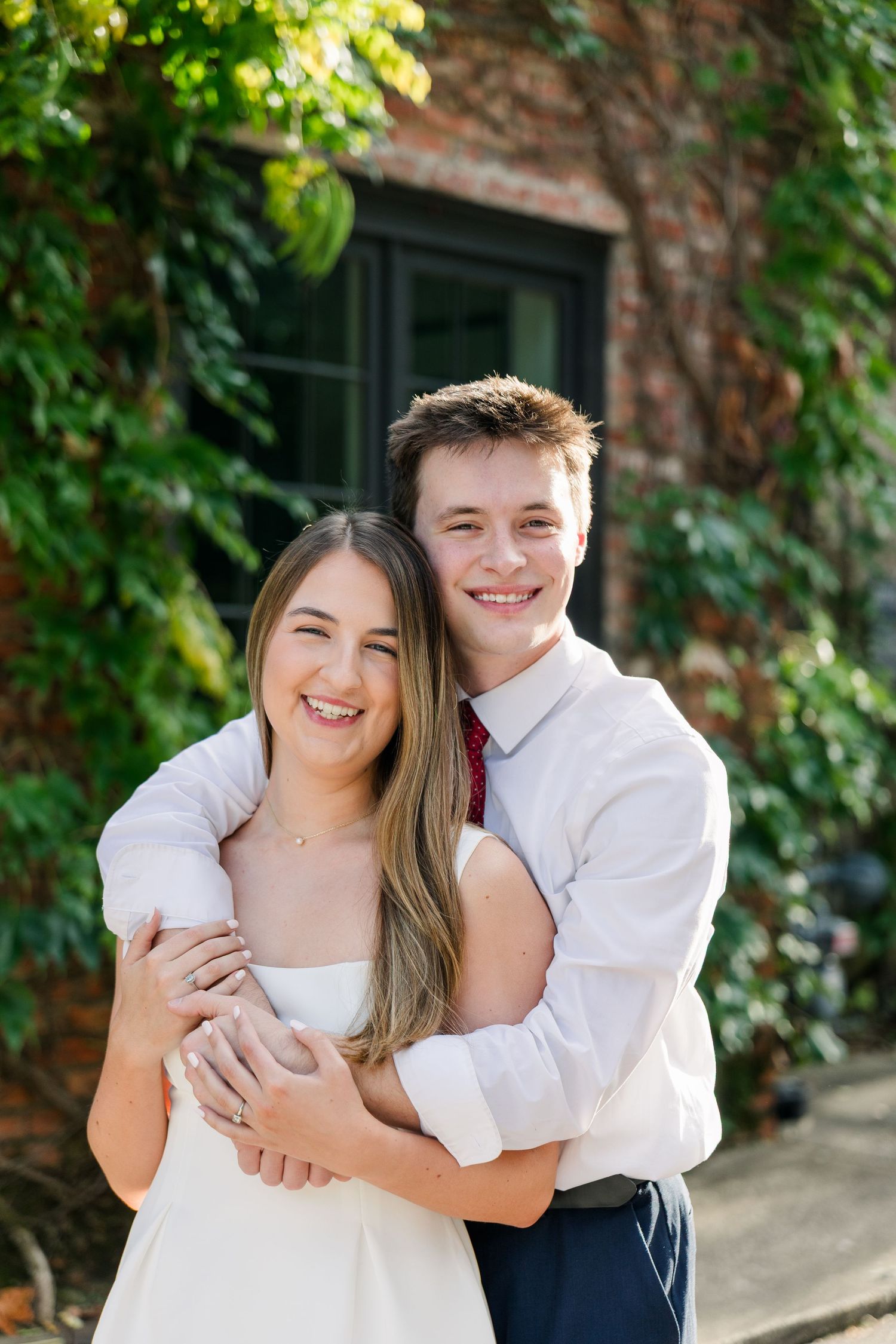 A happy young couple hugs in front of a brick building with lush green ivy.