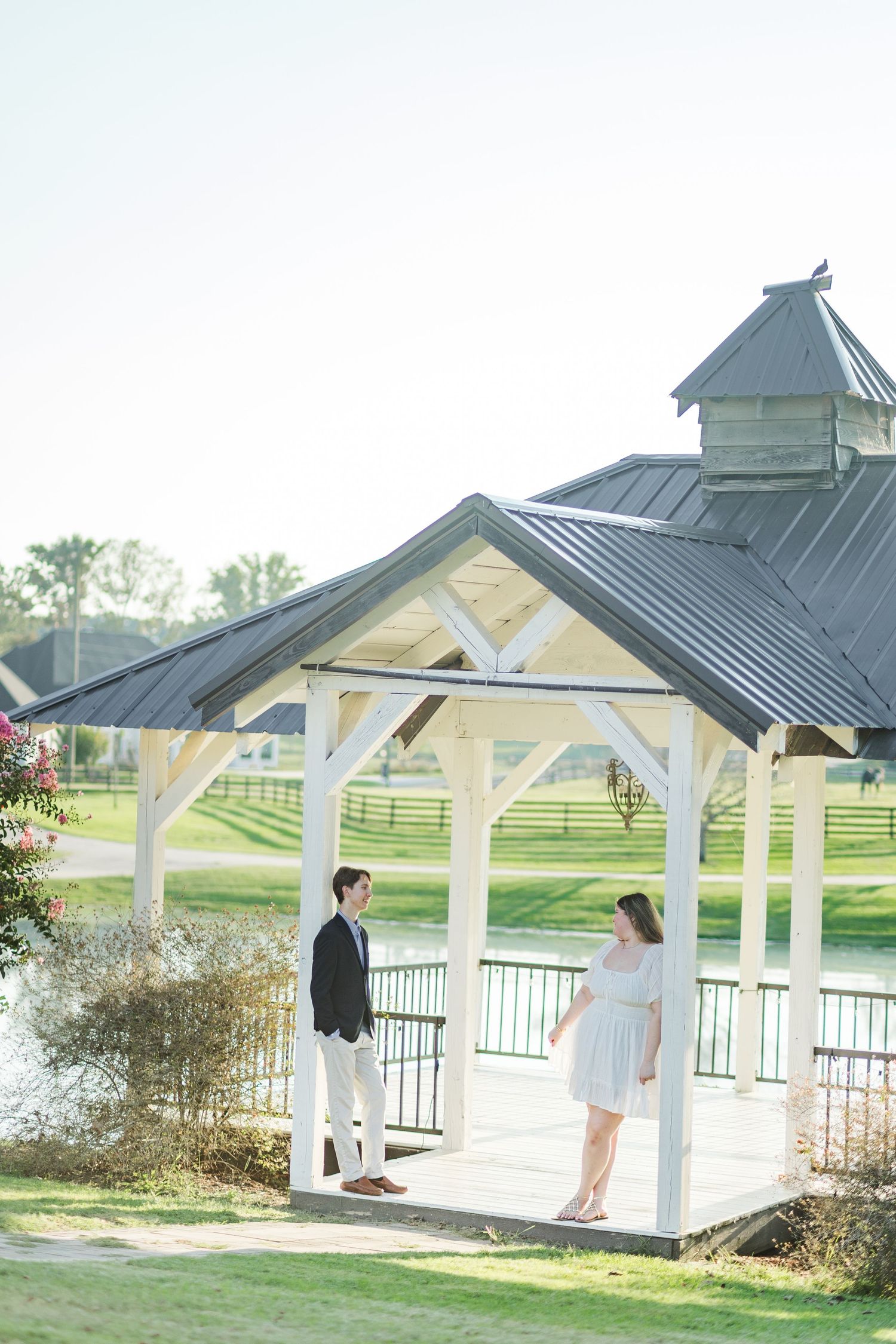Couple stands facing each other in a white gazebo by a lake, surrounded by lush greenery on a sunny day.