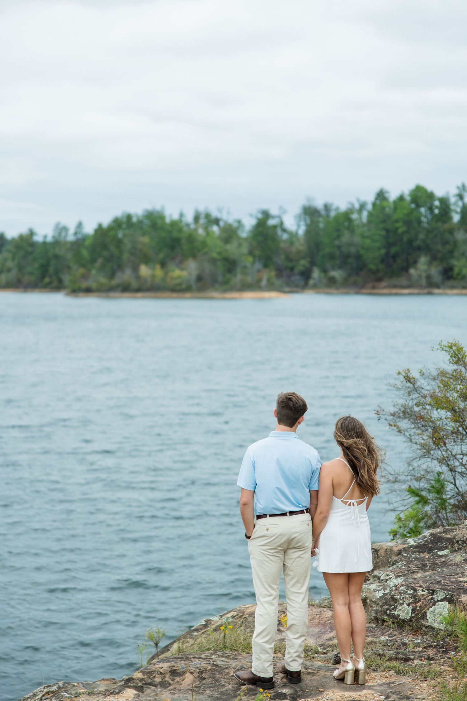 Couple standing close together on a rocky shoreline, looking out over a calm lake toward a tree-lined island.