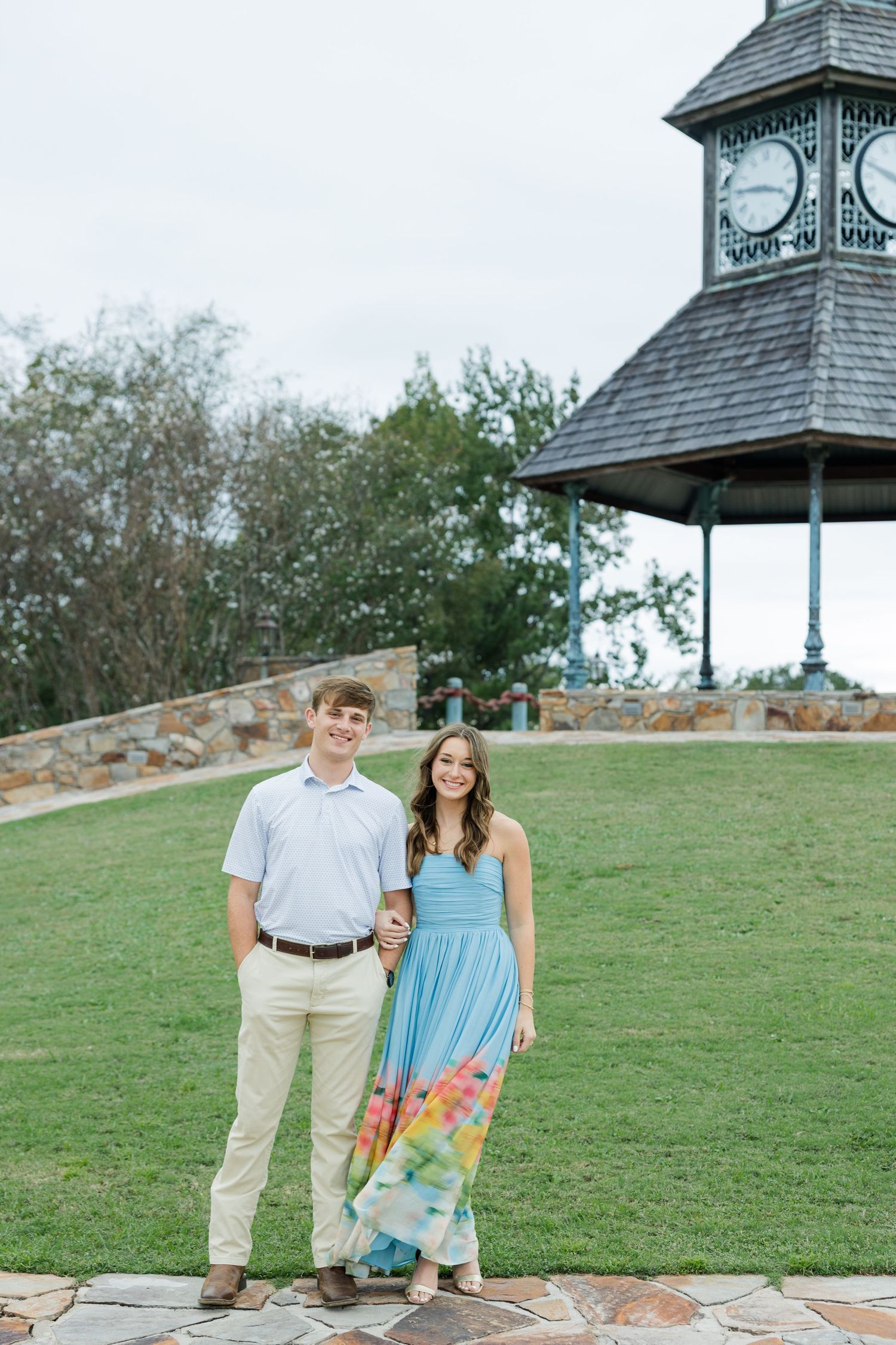 Smiling couple holding hands on a stone path in front of a grassy hill and a clock tower gazebo during an engagement photo session.
