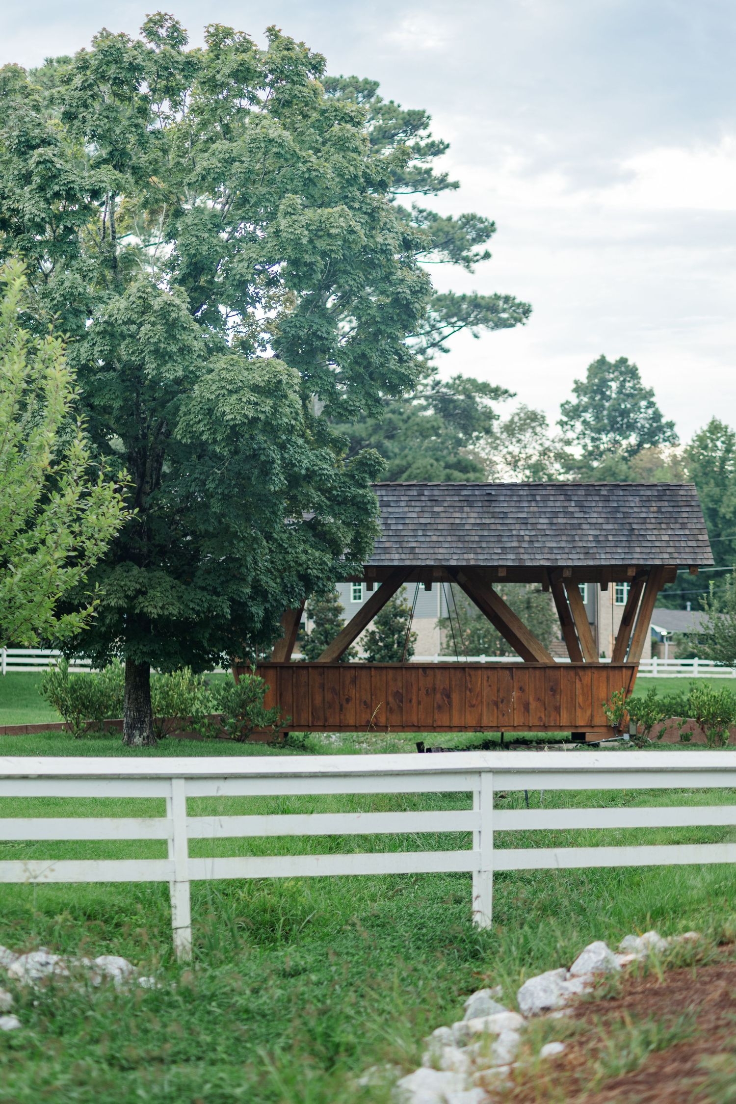 Rustic wooden gazebo with dark shingled roof surrounded by lush green trees and white fence on a farm property.