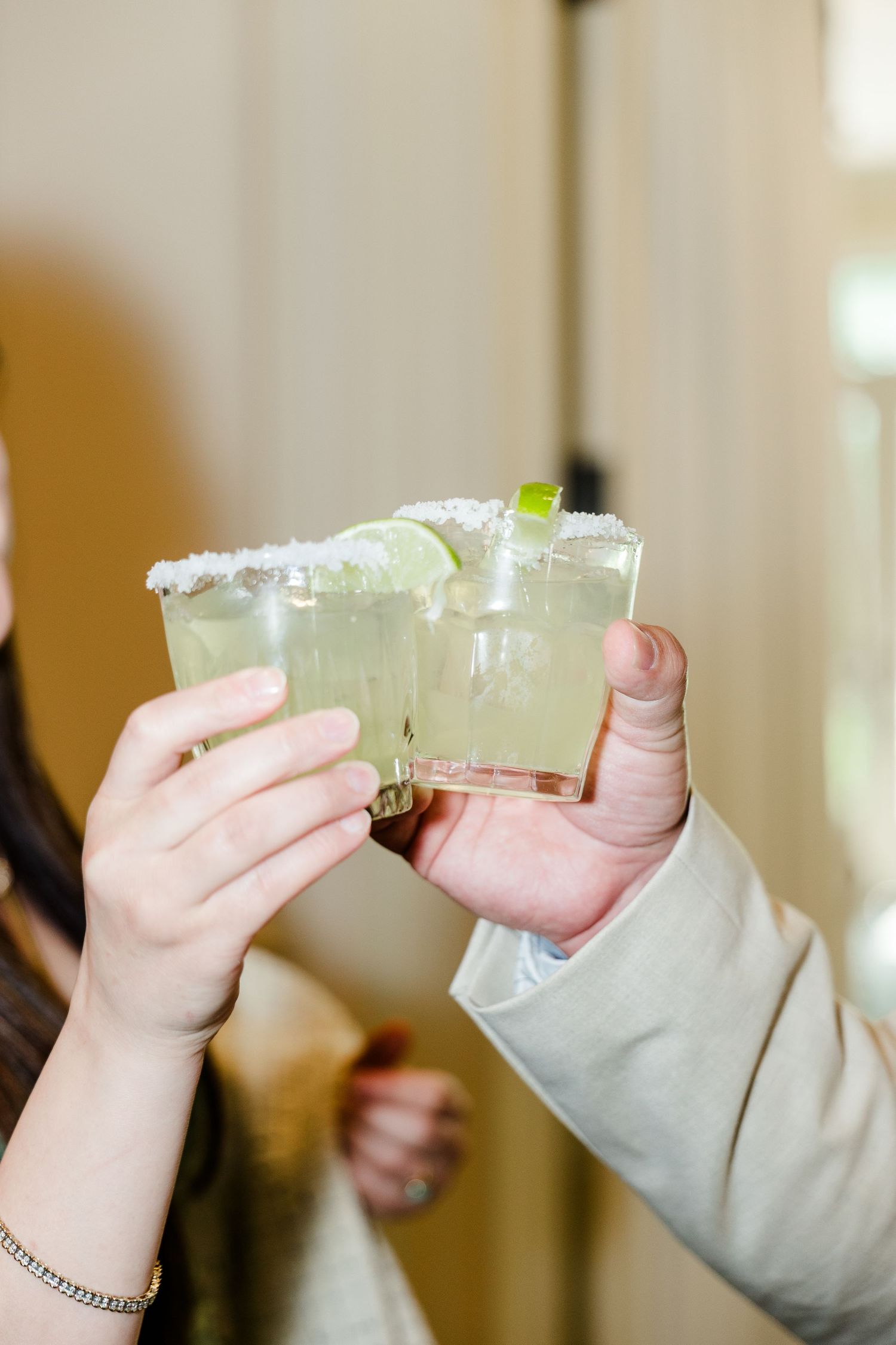 Two hands clinking salt-rimmed margarita glasses with lime garnish at a celebration.