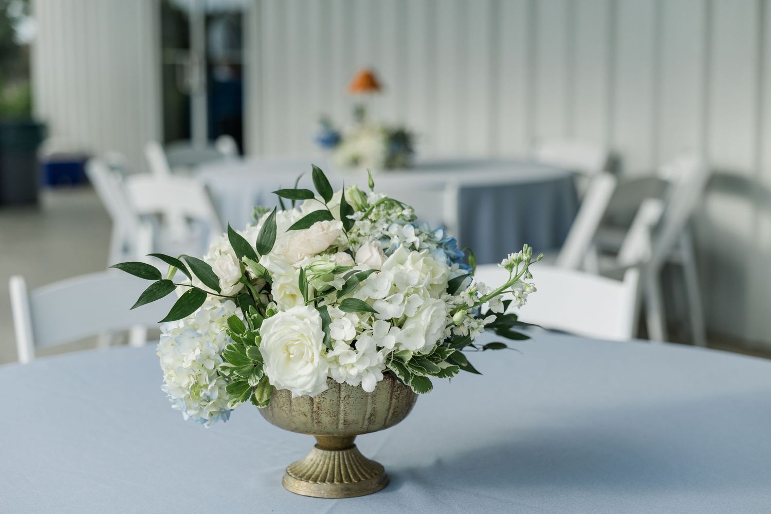 Gold urn centerpiece filled with white roses, hydrangeas, and greenery on blue linen reception table outdoors.