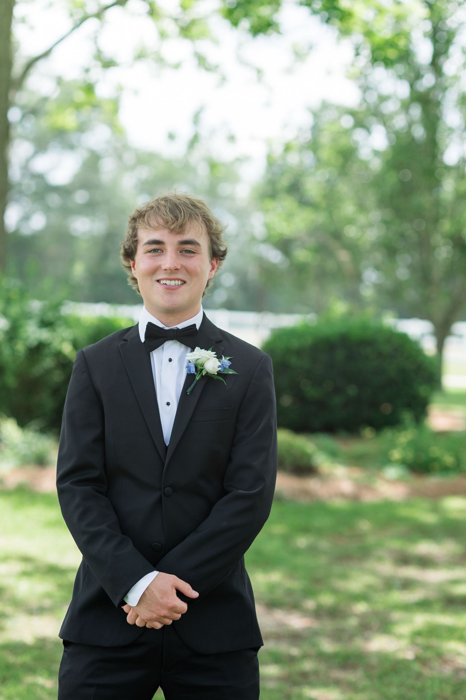 Young groom in black tuxedo with bow tie and boutonniere smiling outdoors at wedding.
