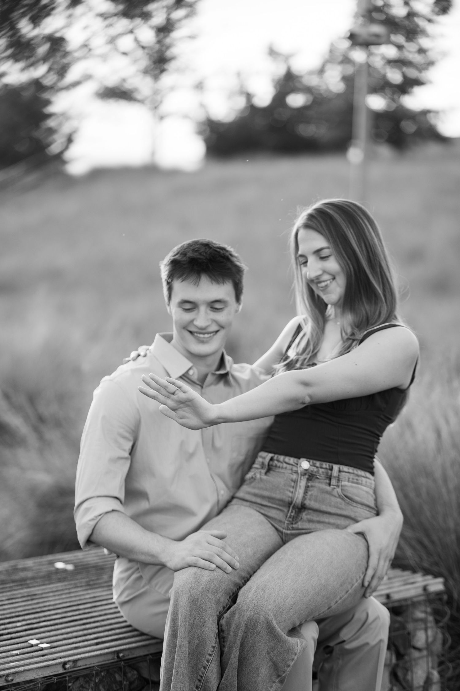 A black and white photo of a laughing couple sitting on a bench in a field at sunset.