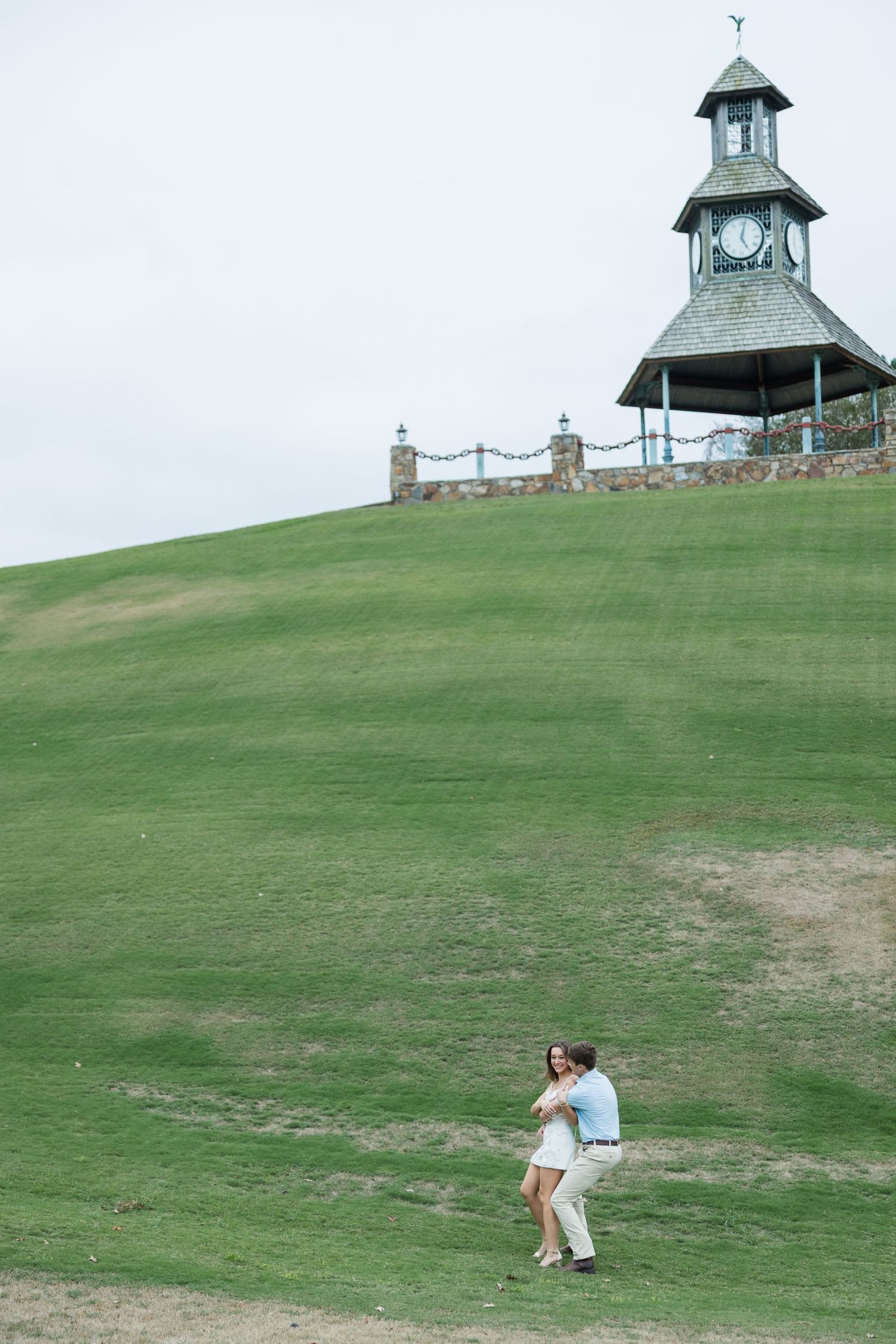 Couple embraces on a large green grassy hillside below a clock tower pavilion with a stone wall under a soft, overcast sky.