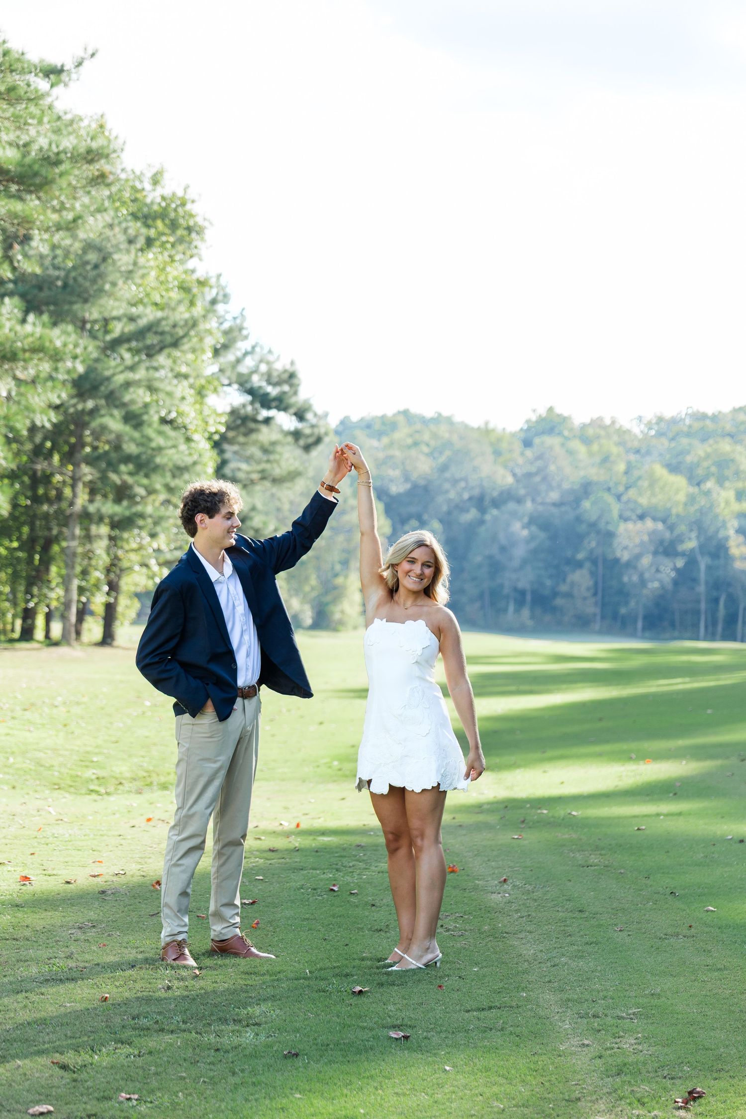 Couple dancing outdoors on green golf course, man in navy blazer twirling woman in white mini dress among trees.