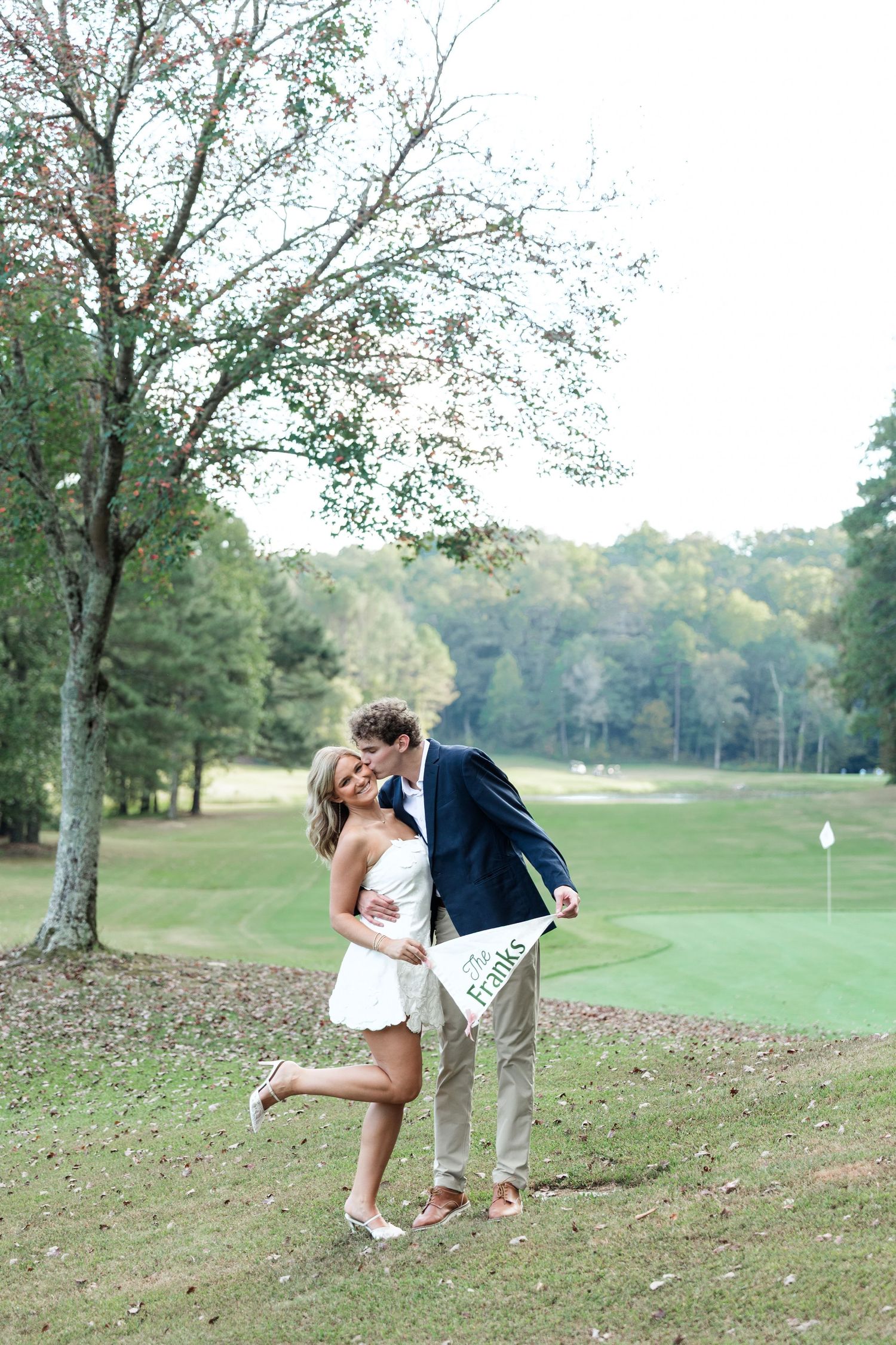 Couple kissing on golf course, woman holding pennant flag, with fairway and trees behind.