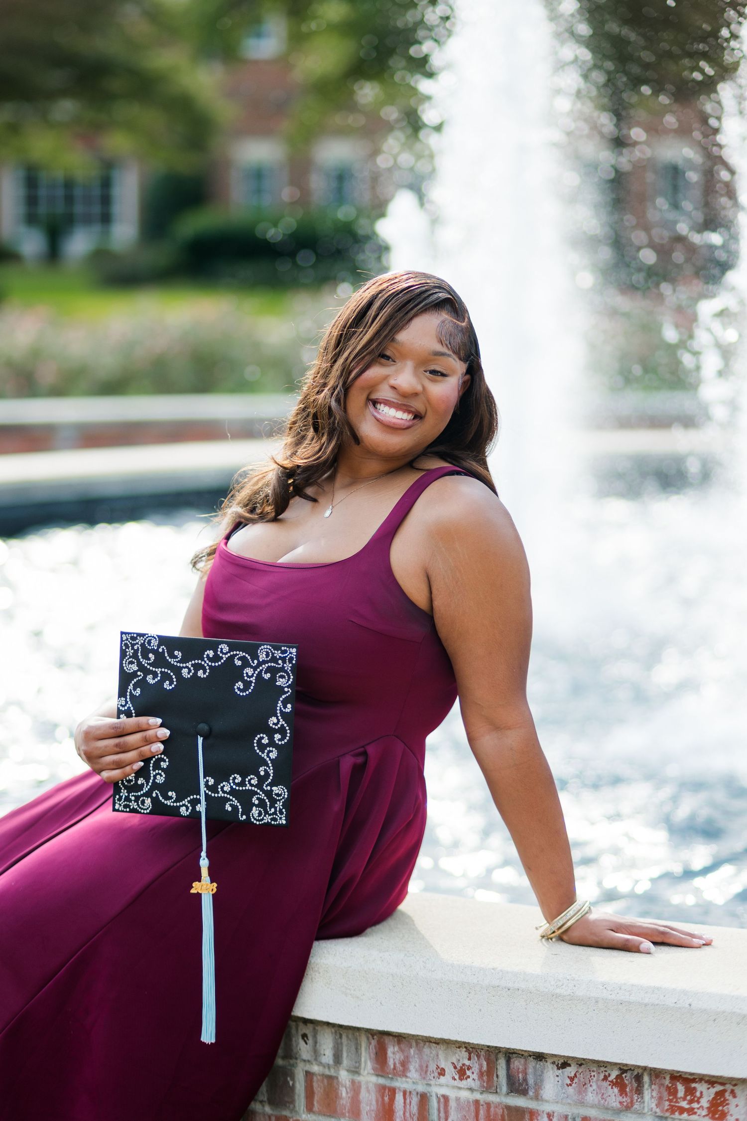 Smiling graduate in burgundy dress holds decorated cap by fountain on college campus.